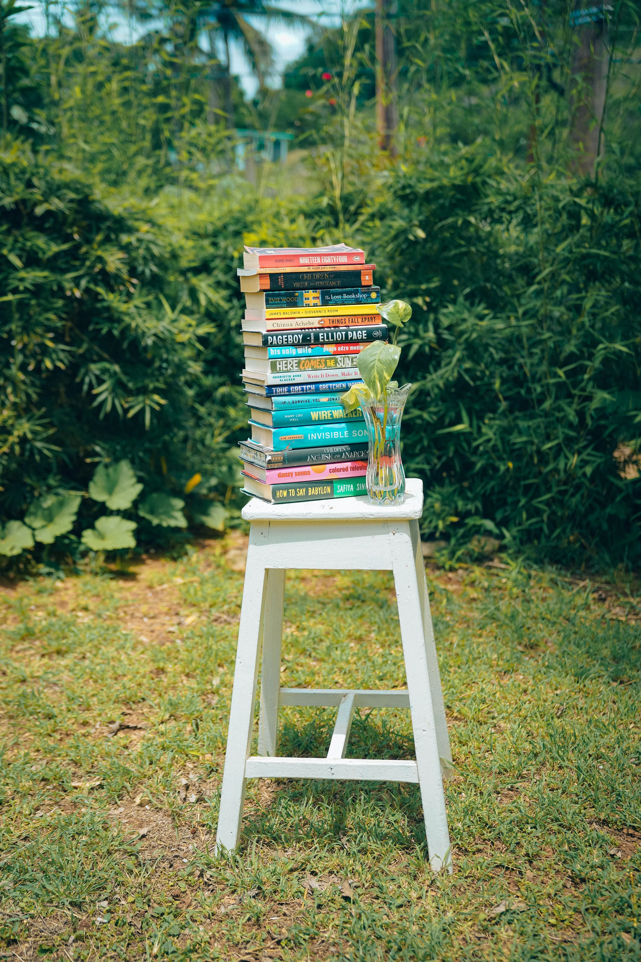 A white wooden stool outdoors on grass, with a tall stack of colorful books and a glass vase with green leaves in front of a green garden background.