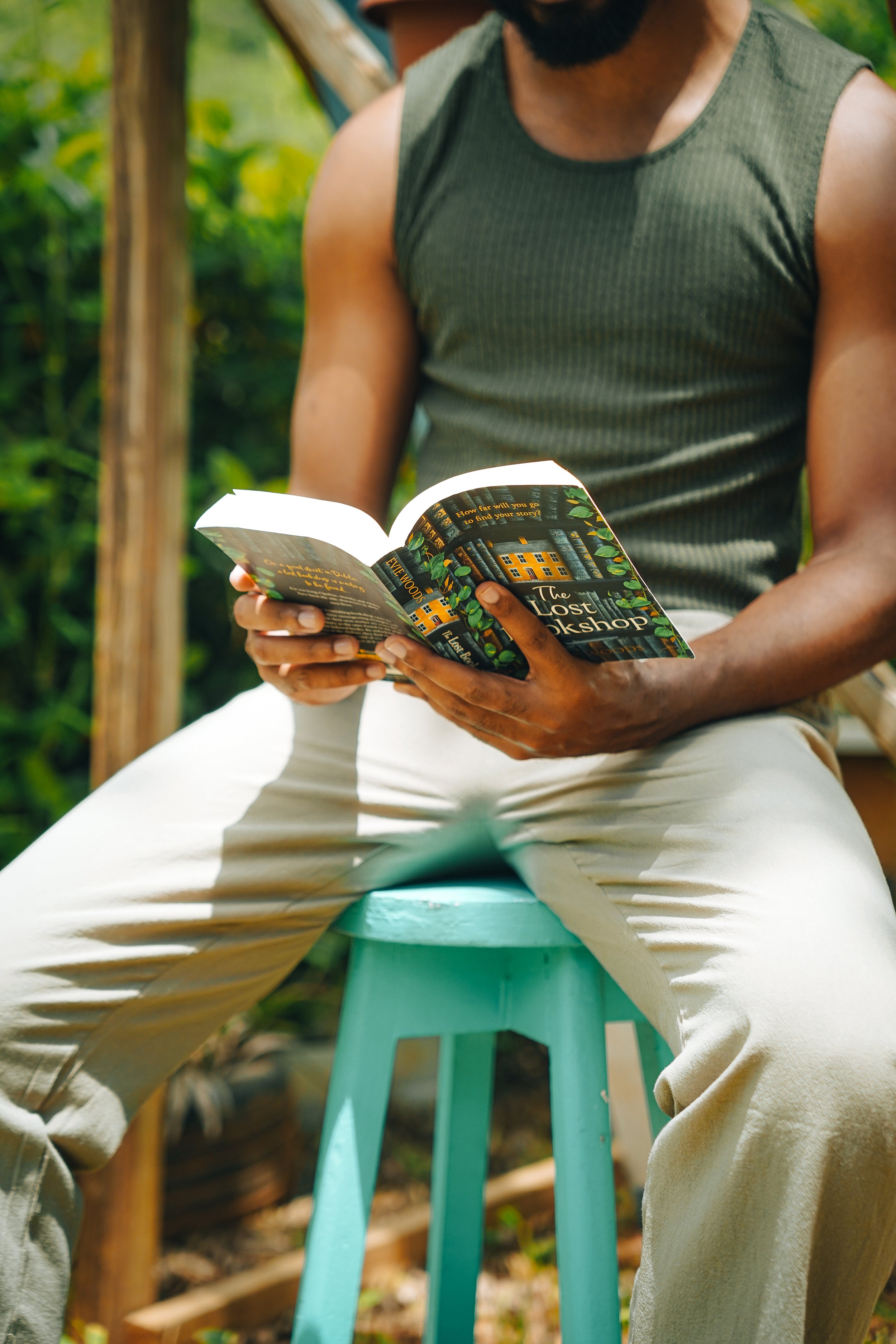 A person sitting on a turquoise stool outdoors, reading a book titled 'The Lost Workshop', wearing a sleeveless green shirt and beige pants, with green foliage in the background.