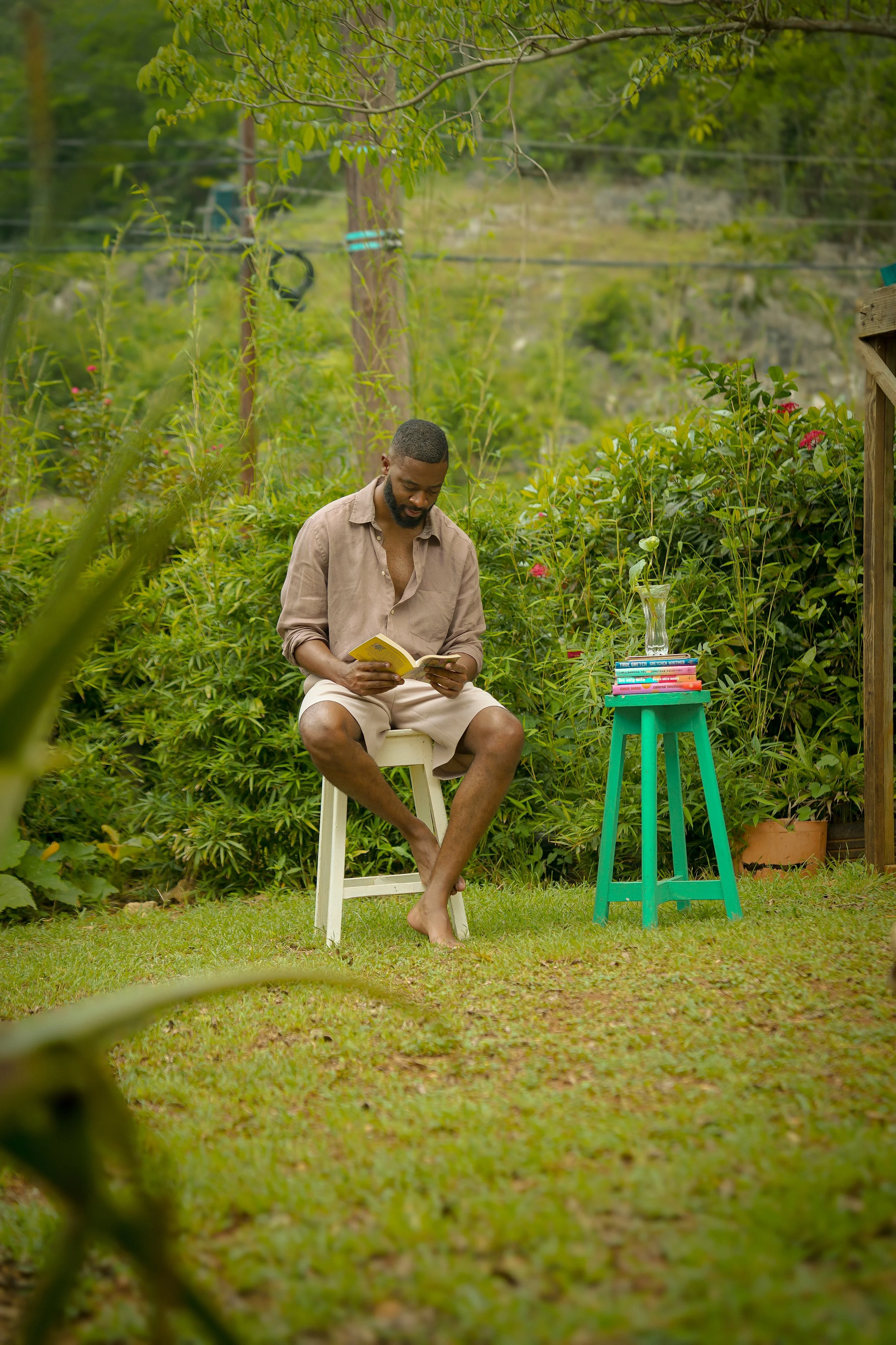 A man sitting outside on a white stool reading a book. There is a small teal table with a stack of books and a flower in a glass vase beside him. The setting is lush and green, with trees and bushes in the background.