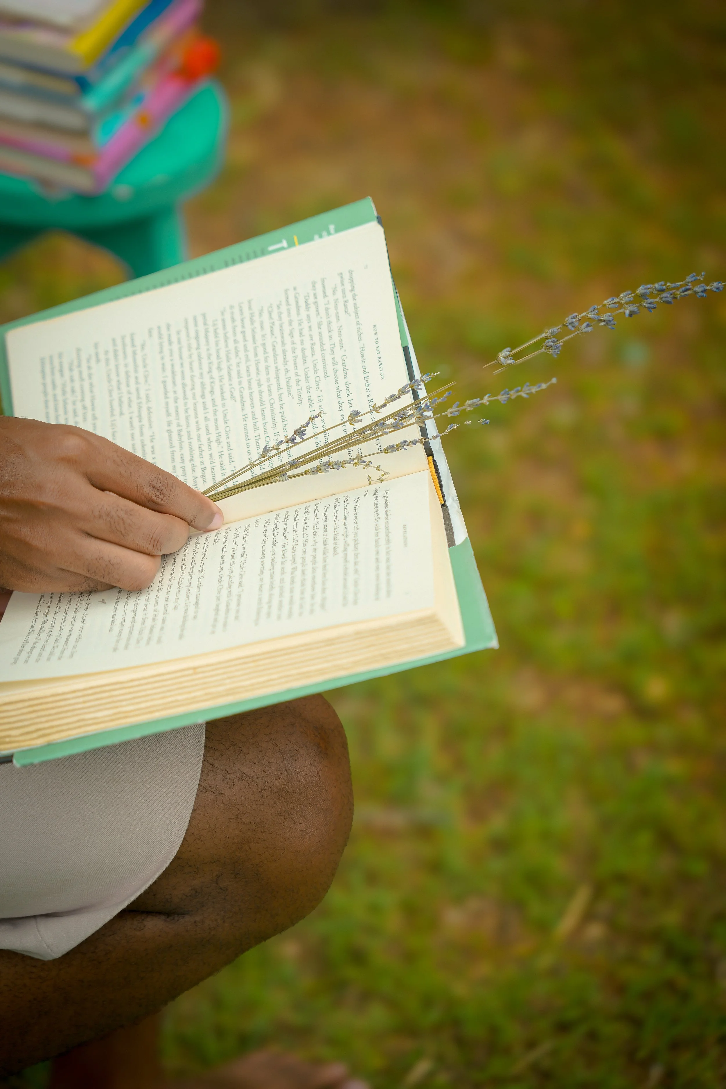 Person reading a book outdoors, holding a sprig of lavender, with a green portable table with books in the background, and grass on the ground.