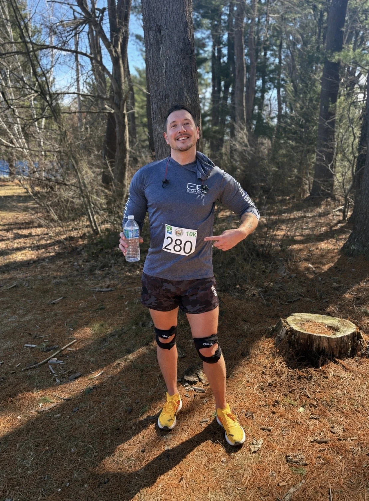 A man standing in a wooded area during a running event, holding a water bottle, wearing race bib number 280, athletic shorts, knee braces, yellow running shoes, and a long-sleeve shirt with event or team logos, smiling and pointing at his bib.