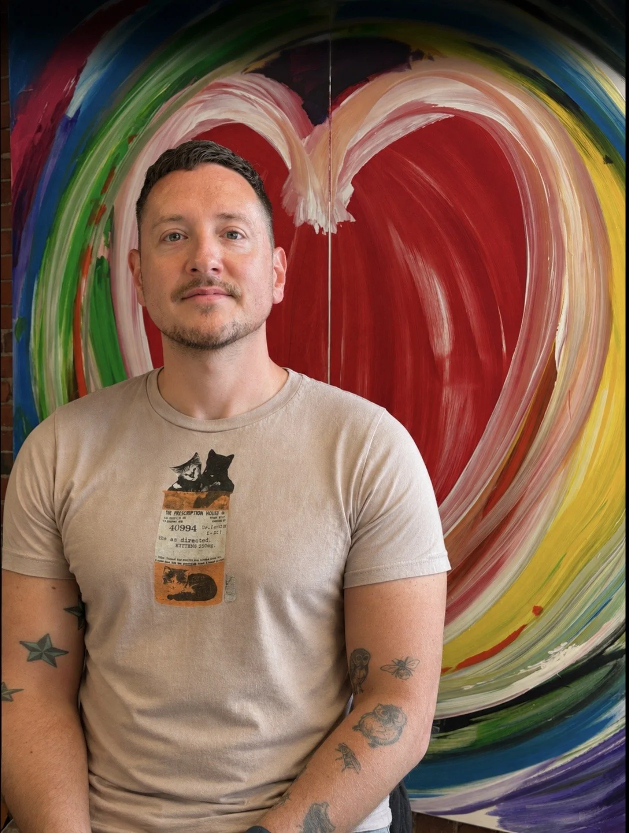 A man with short dark hair, tattoos on his arms, and a beige t-shirt with a graphic of cats and a ticket stub design standing in front of a colorful abstract painting with a large red heart at its center.