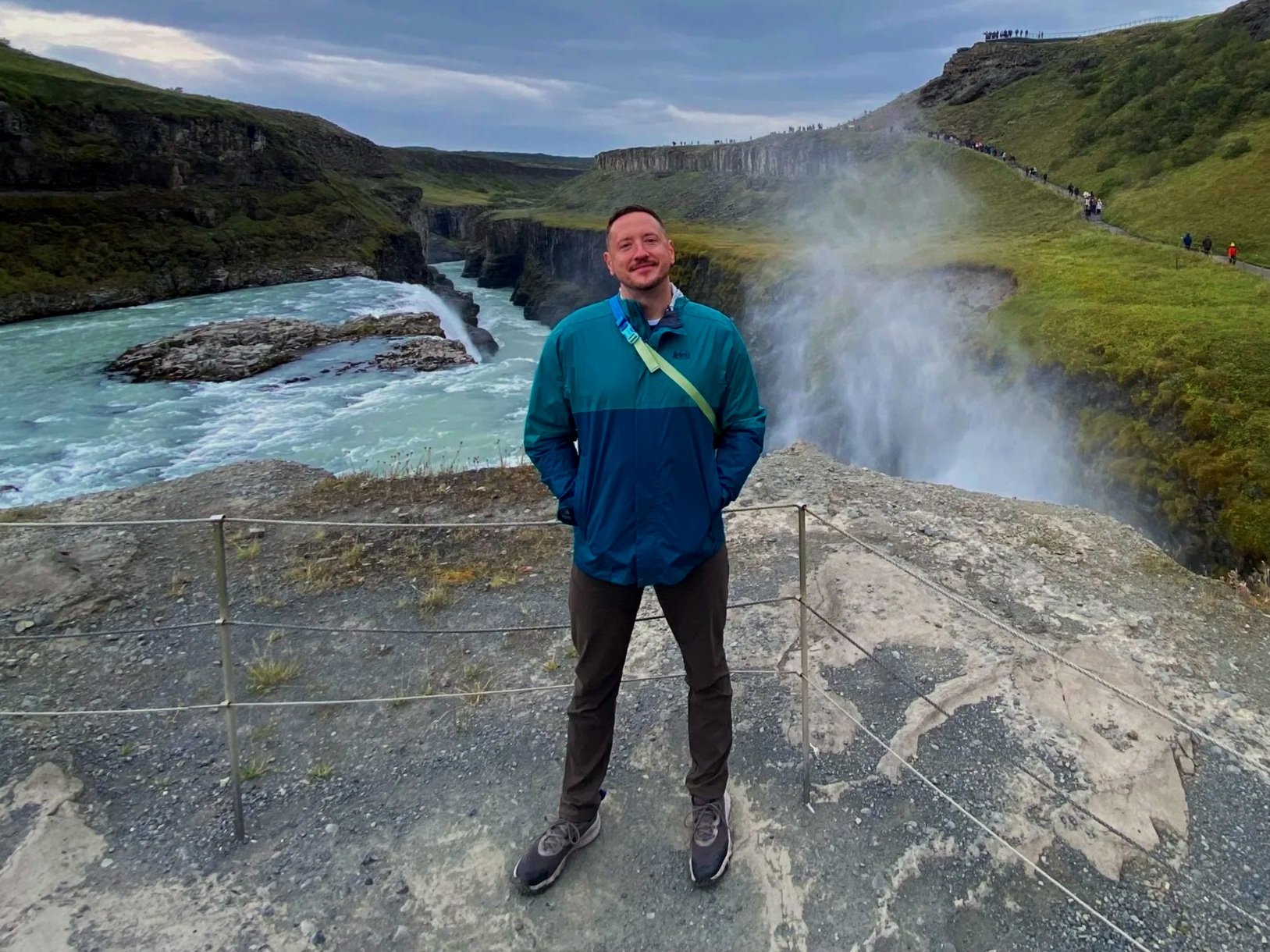 A man standing outdoors in front of a waterfall and river with green hills and cliffs, wearing a blue jacket and black pants.