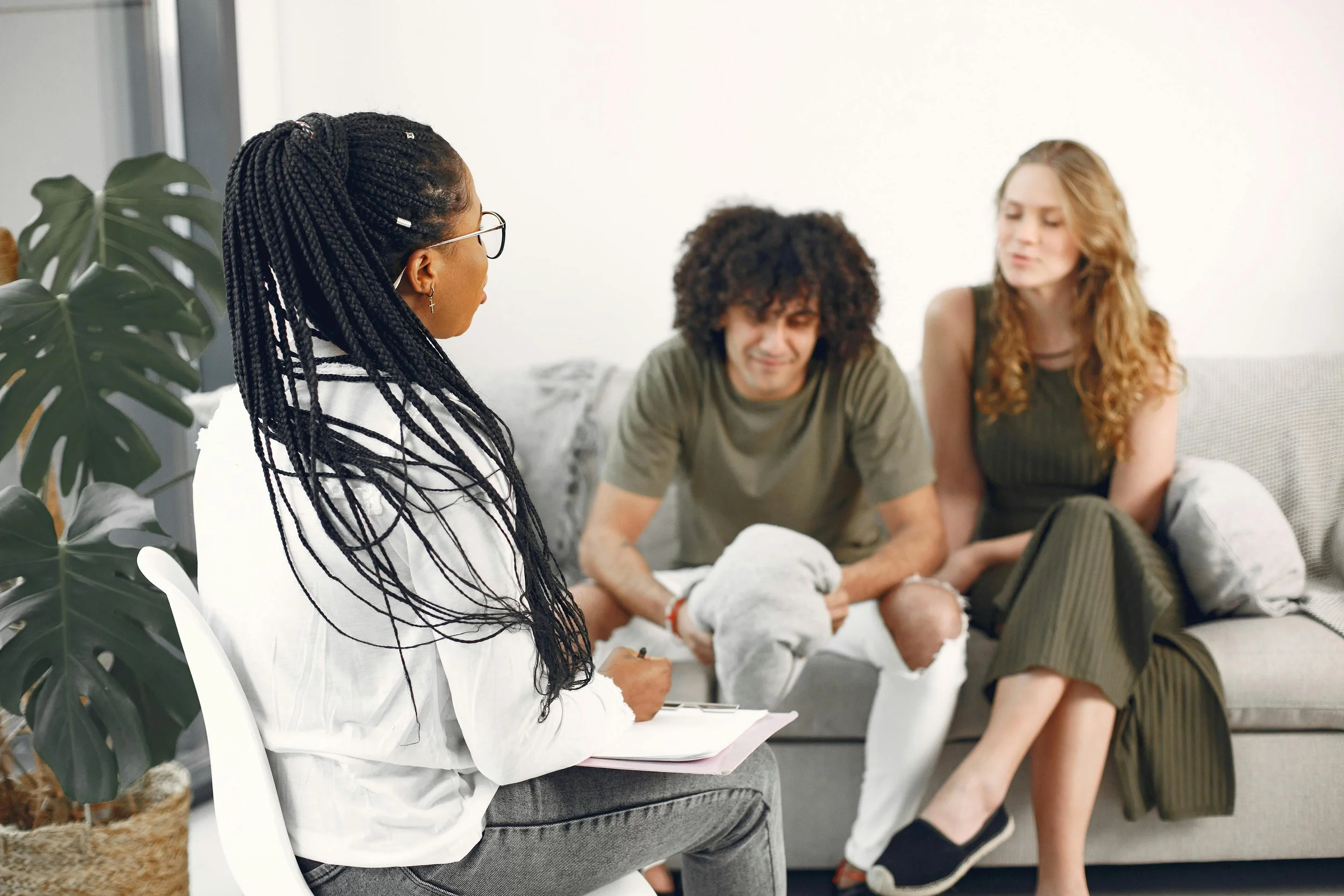 A diverse group of young adults sitting on a sofa in a living room, engaged in conversation. The group includes a woman with braided hair, a man with curly hair, and a woman with long wavy hair. There is a large plant in the foreground.