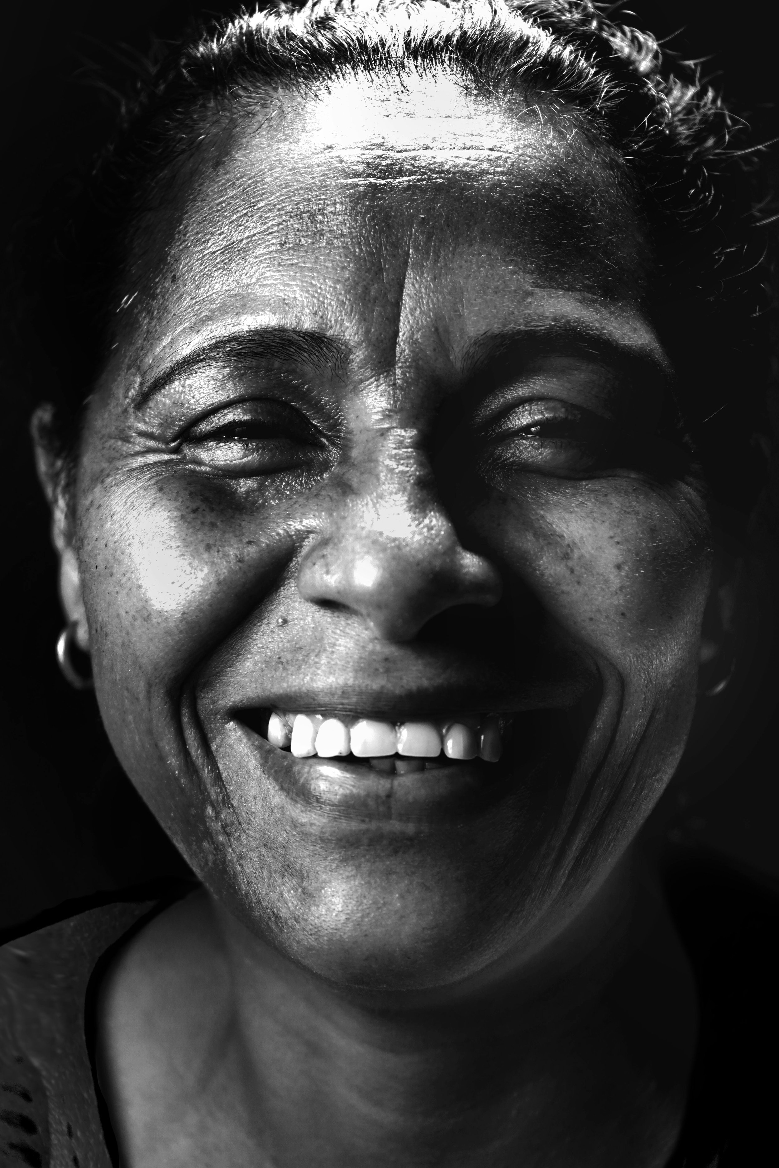 Close-up of a smiling woman with dark skin, curly hair pulled back, and wearing earrings, captured in black and white.
