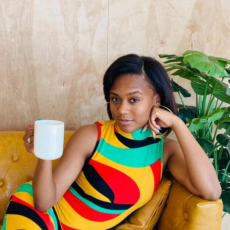 A woman with medium skin tone and black hair in a side part, wearing hoop earrings and a colorful sleeveless dress, sitting on a yellow sofa with a wooden background and a large green houseplant beside her, holding a white mug.