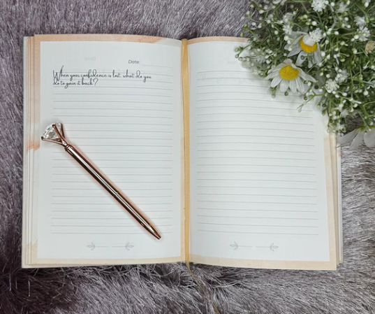An open journal with lined pages lying on a grey furry surface, with a rose gold pen resting on the left page and a bouquet of white daisies and small white flowers on the top right corner.
