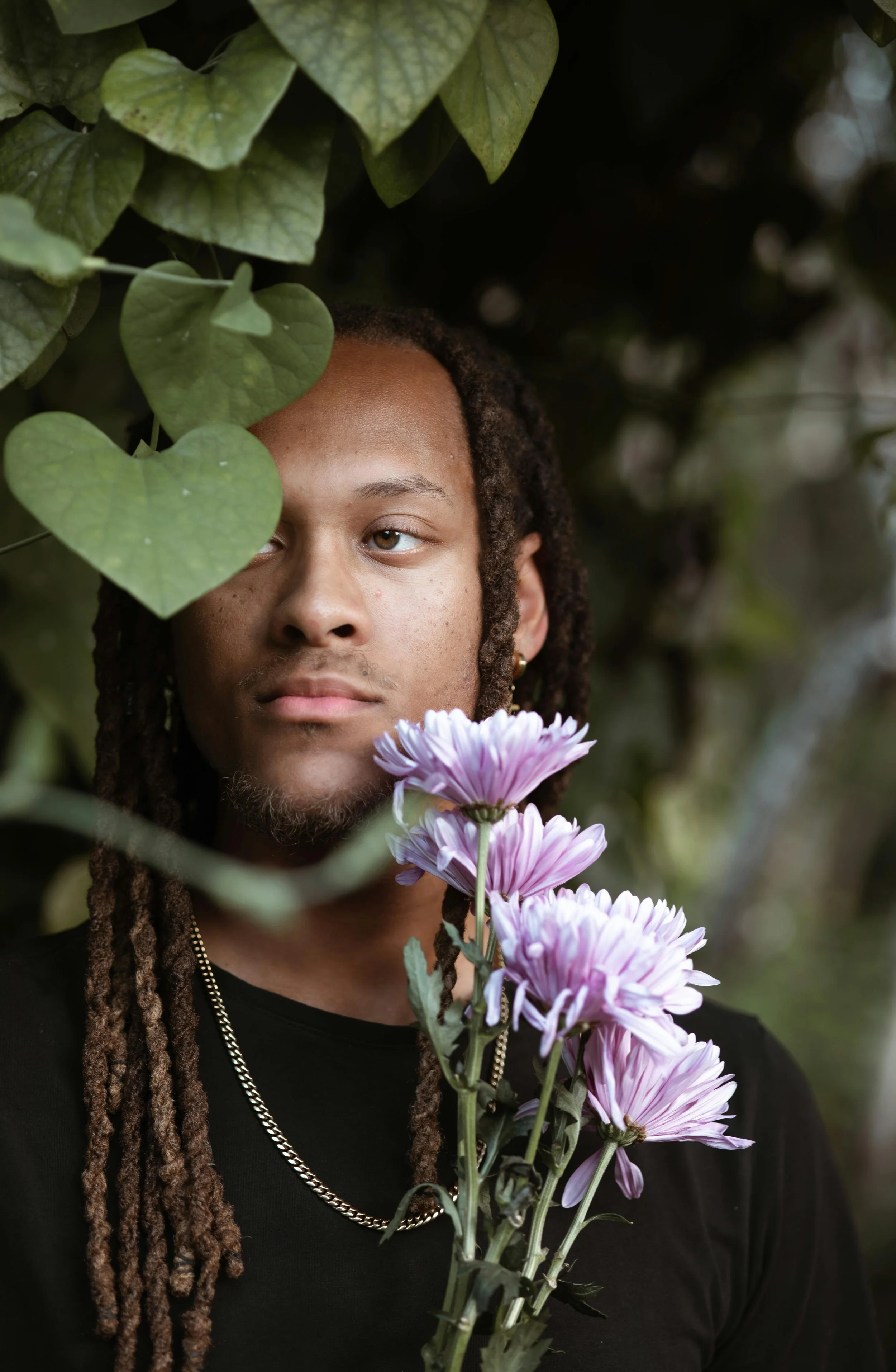 A person with long dreadlocks holding a bunch of pink and purple flowers, partially obscured by green leaves, in an outdoor setting.