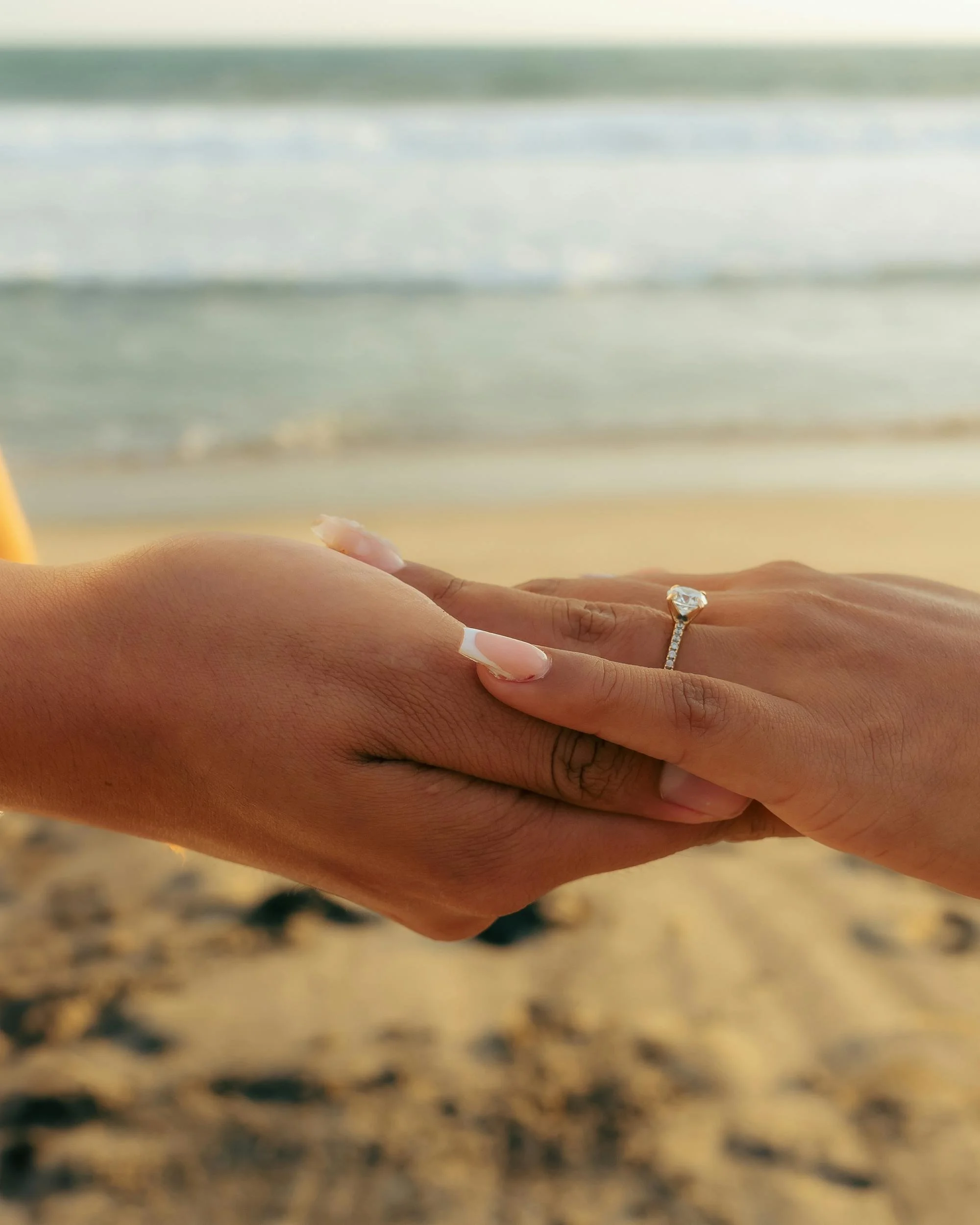 Close-up of two hands holding each other on a beach, one hand wearing an engagement ring with a large diamond, blurred shoreline and water in the background.