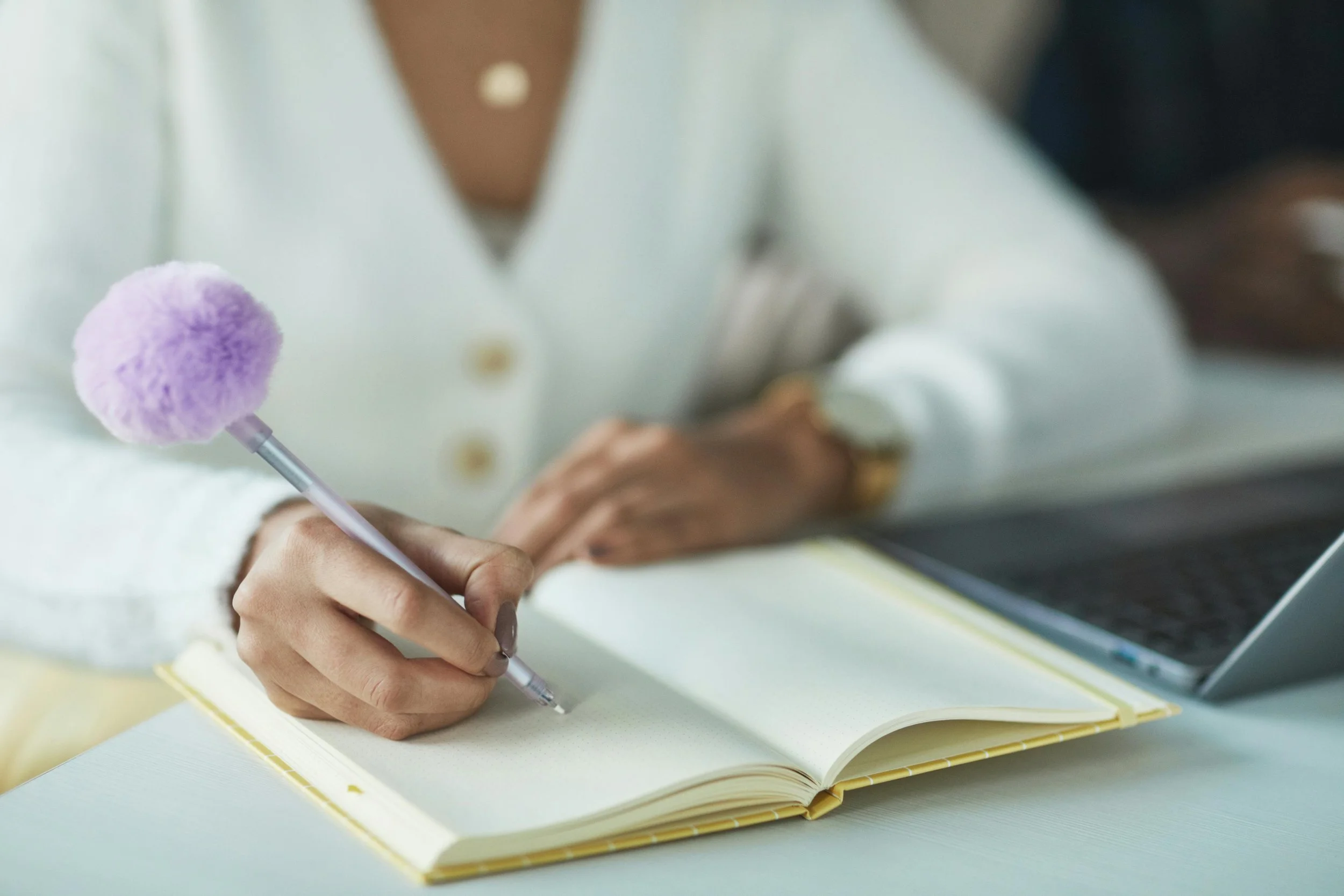 Close-up of a person writing in a notebook with a pen that has a purple fluffy pom-pom on top. The person is wearing a light-colored shirt and has a ring on their hand.