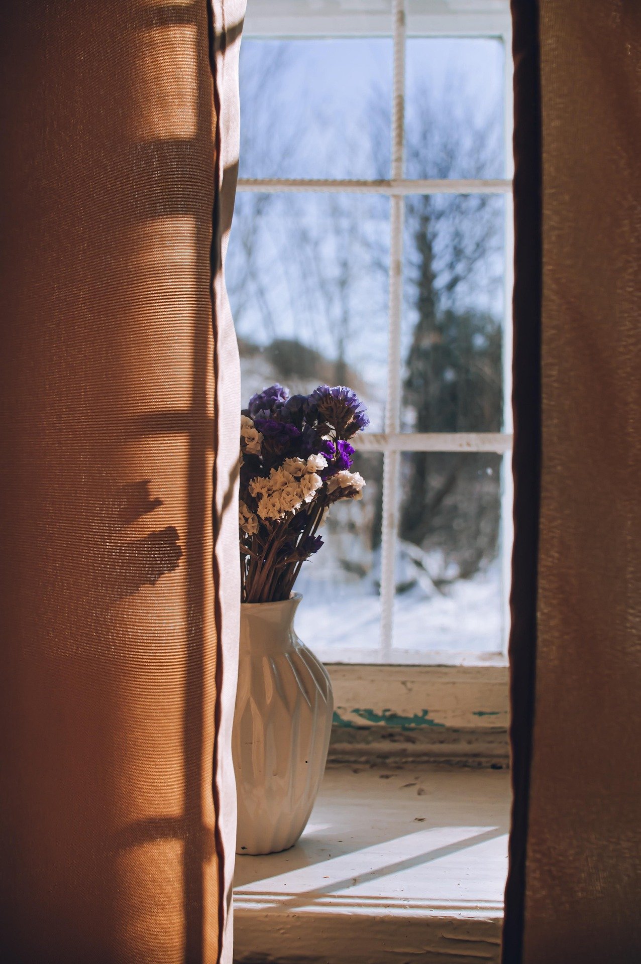 A vase with purple and white flowers on a windowsill, viewed through partly open curtains with sunlight casting shadows.