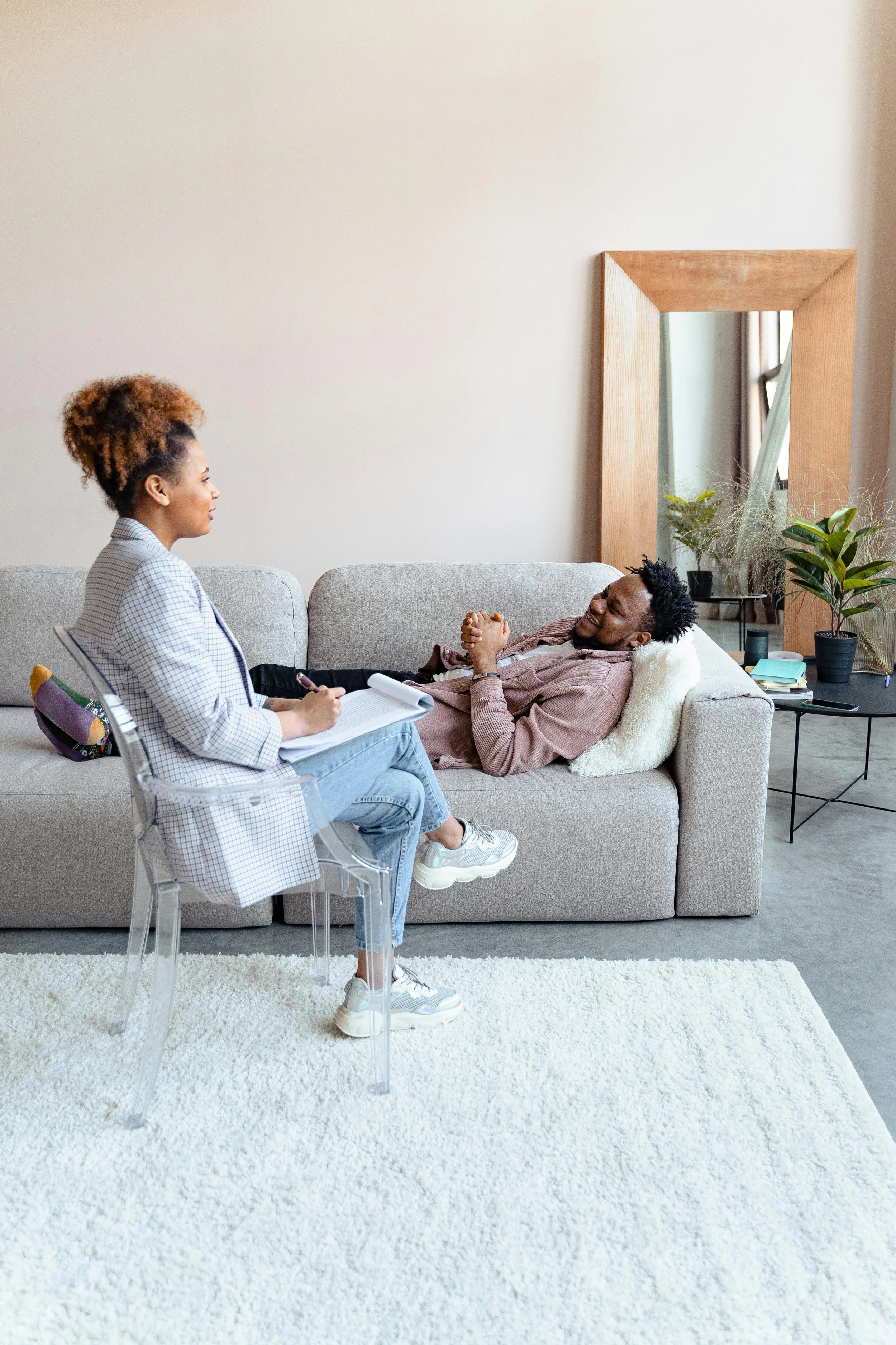 A woman sitting on a transparent chair, taking notes, and talking to a man lying on a light-colored sofa, smiling with his hands clasped. The room has a large mirror, plants, and a beige carpet.