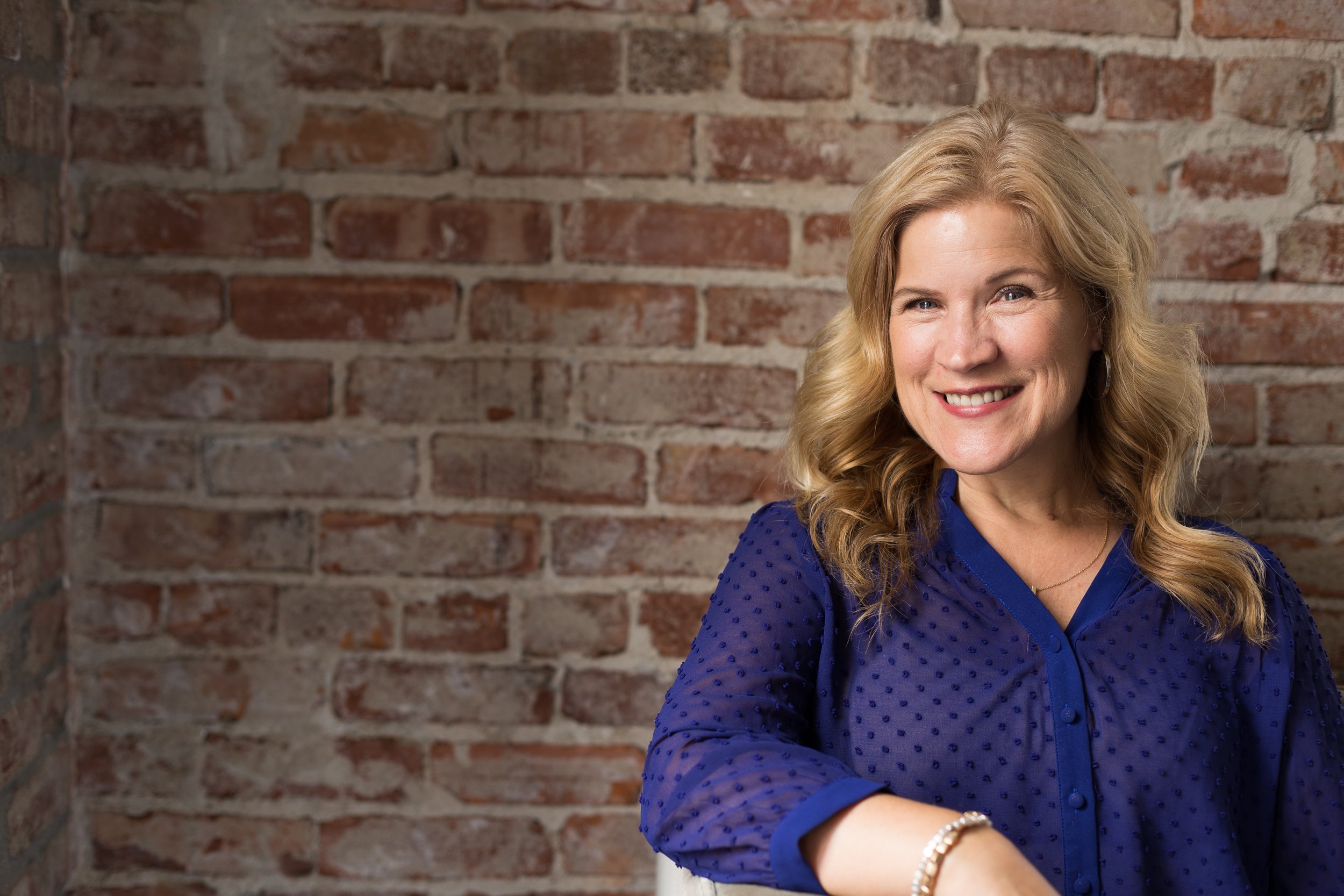 A smiling woman with blonde curly hair, wearing a blue blouse, sitting in front of a brick wall.