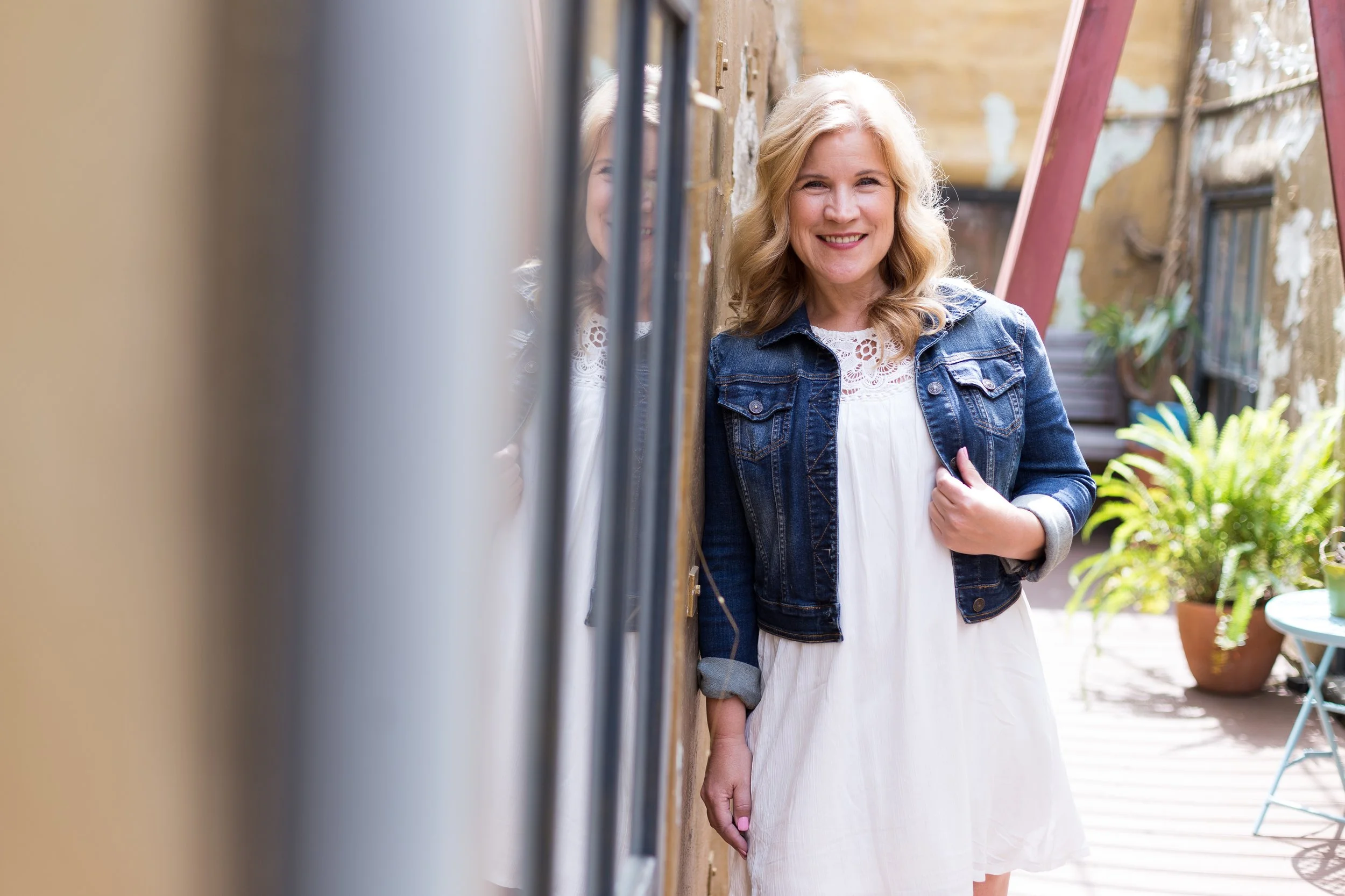 A smiling woman with blonde hair wearing a denim jacket and white dress, standing outdoors next to a brick wall with colorful plants and patio furniture in the background.