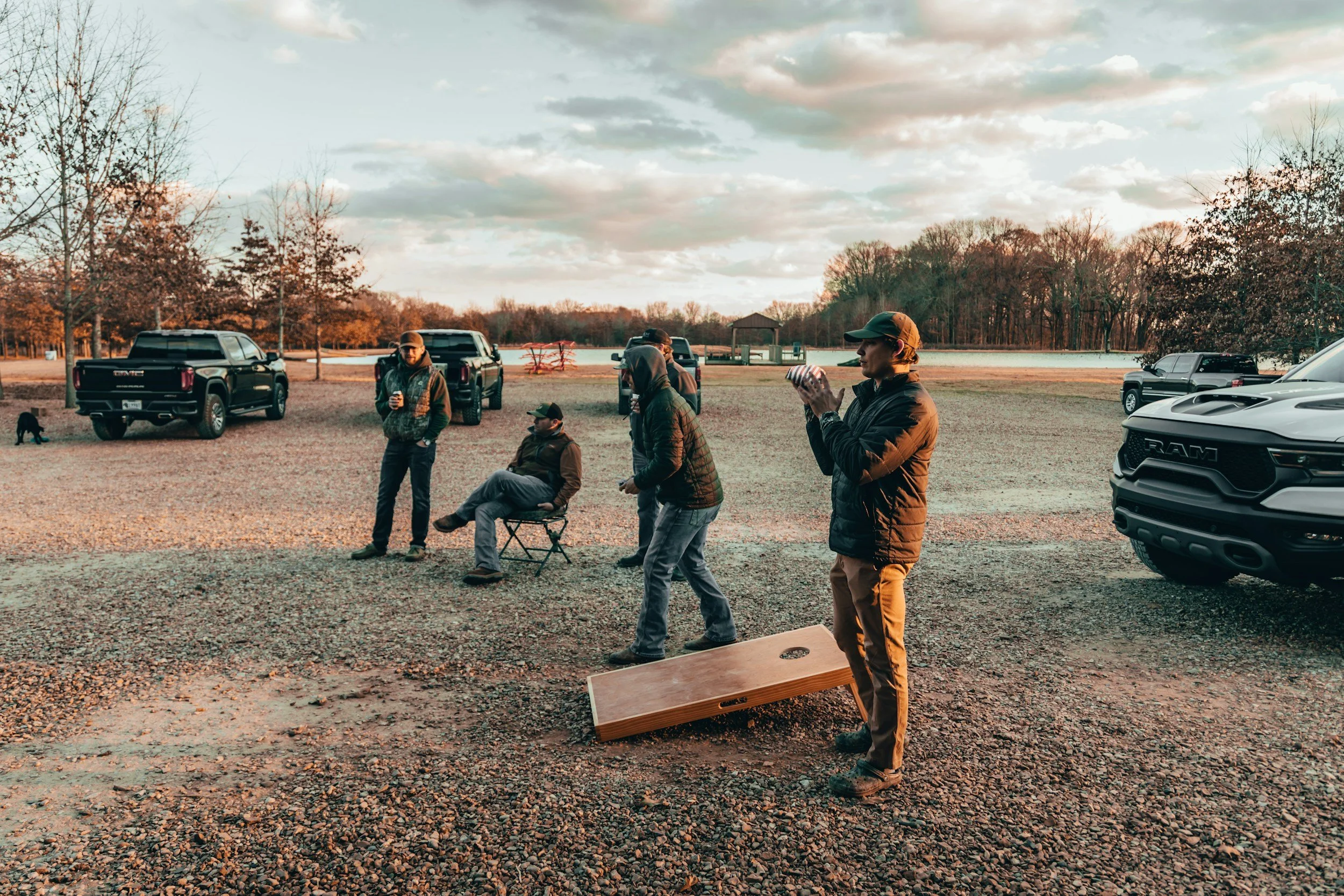 A group of people playing cornhole on a gravel lot near parked trucks and trees during sunset.