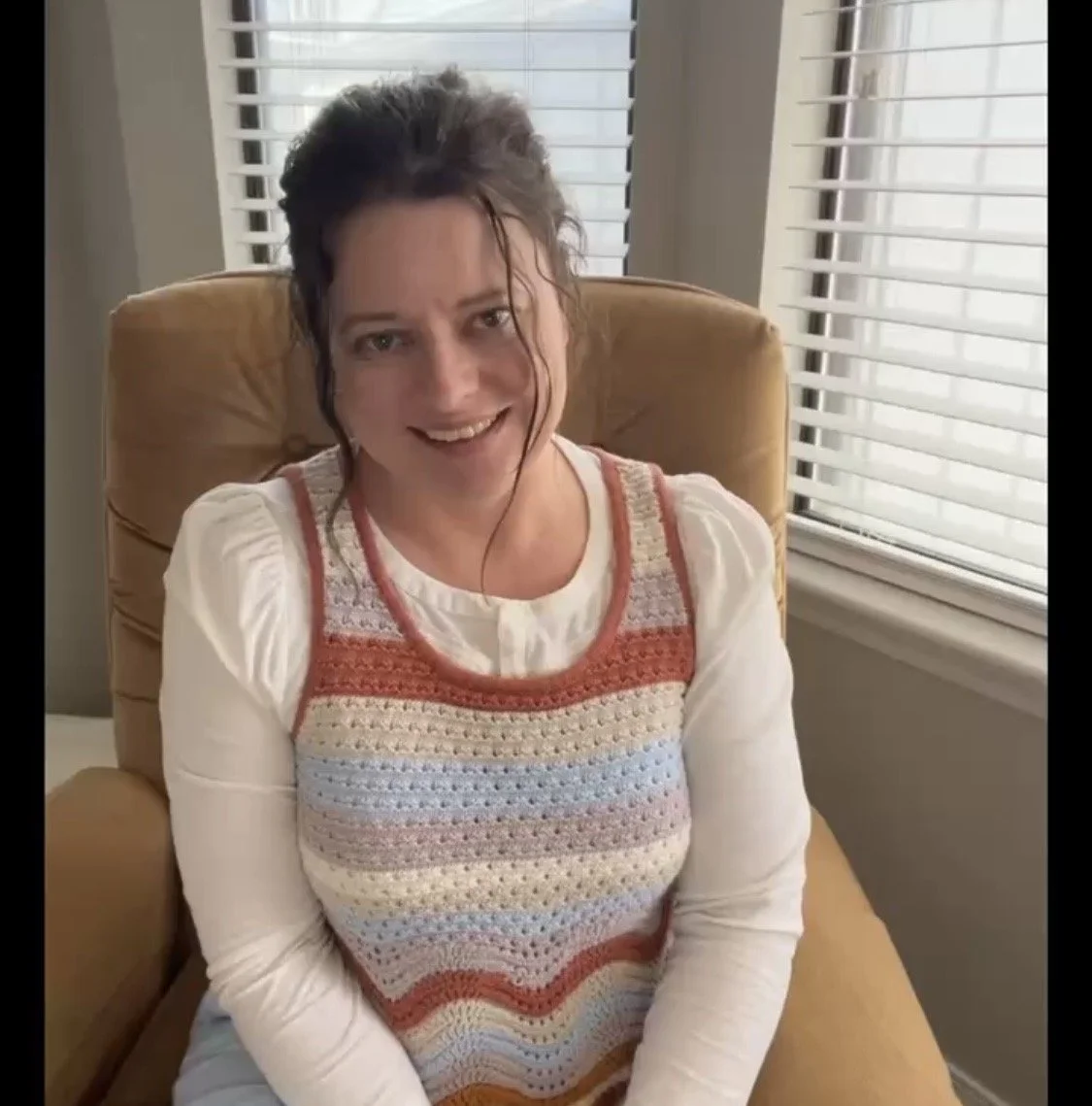 A woman with dark, curly hair smiling while sitting in a tan armchair by a window with white blinds.