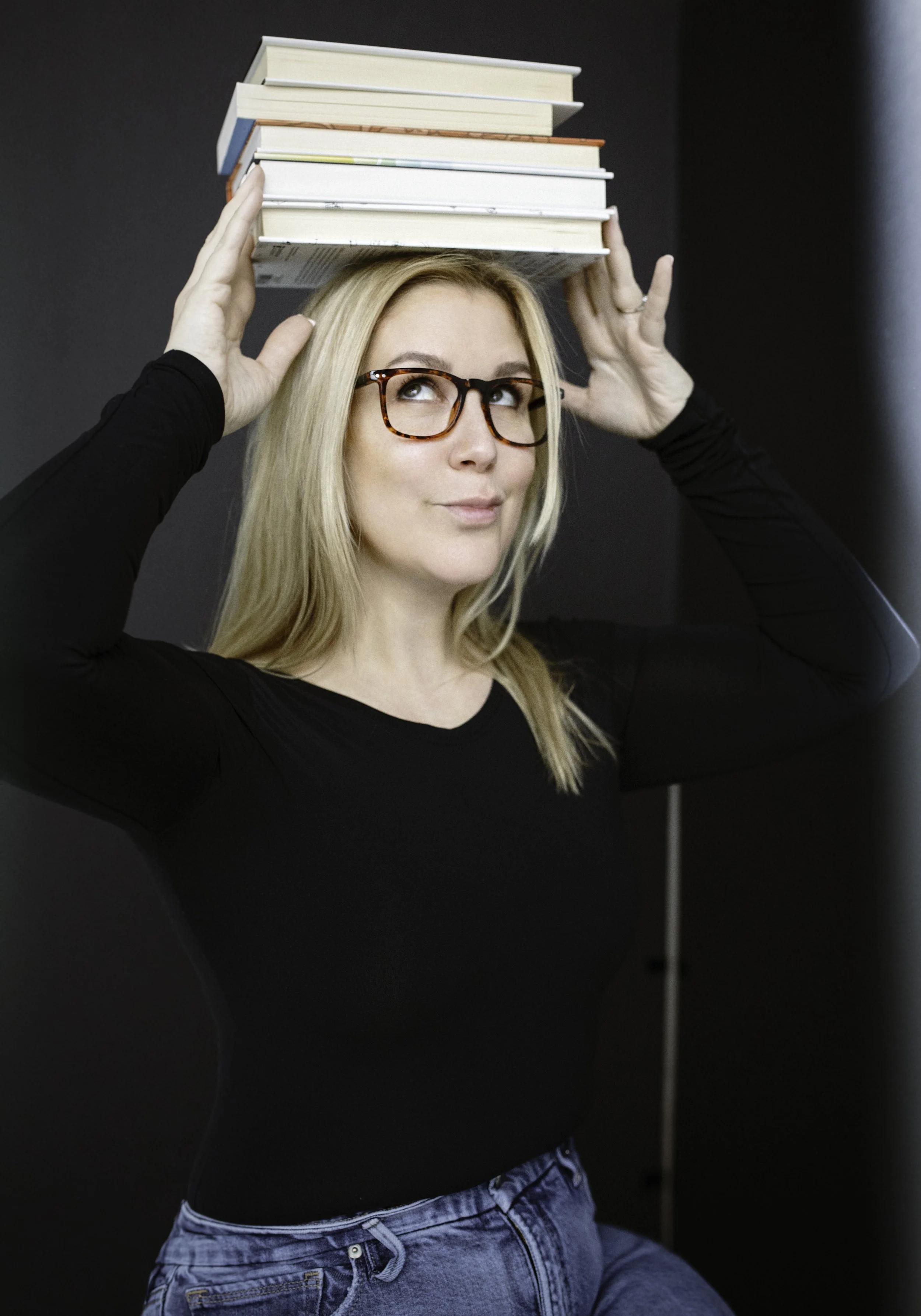 Andrea Mac with blonde hair and glasses balancing a stack of books on her head, wearing a black top and blue jeans against a dark background.