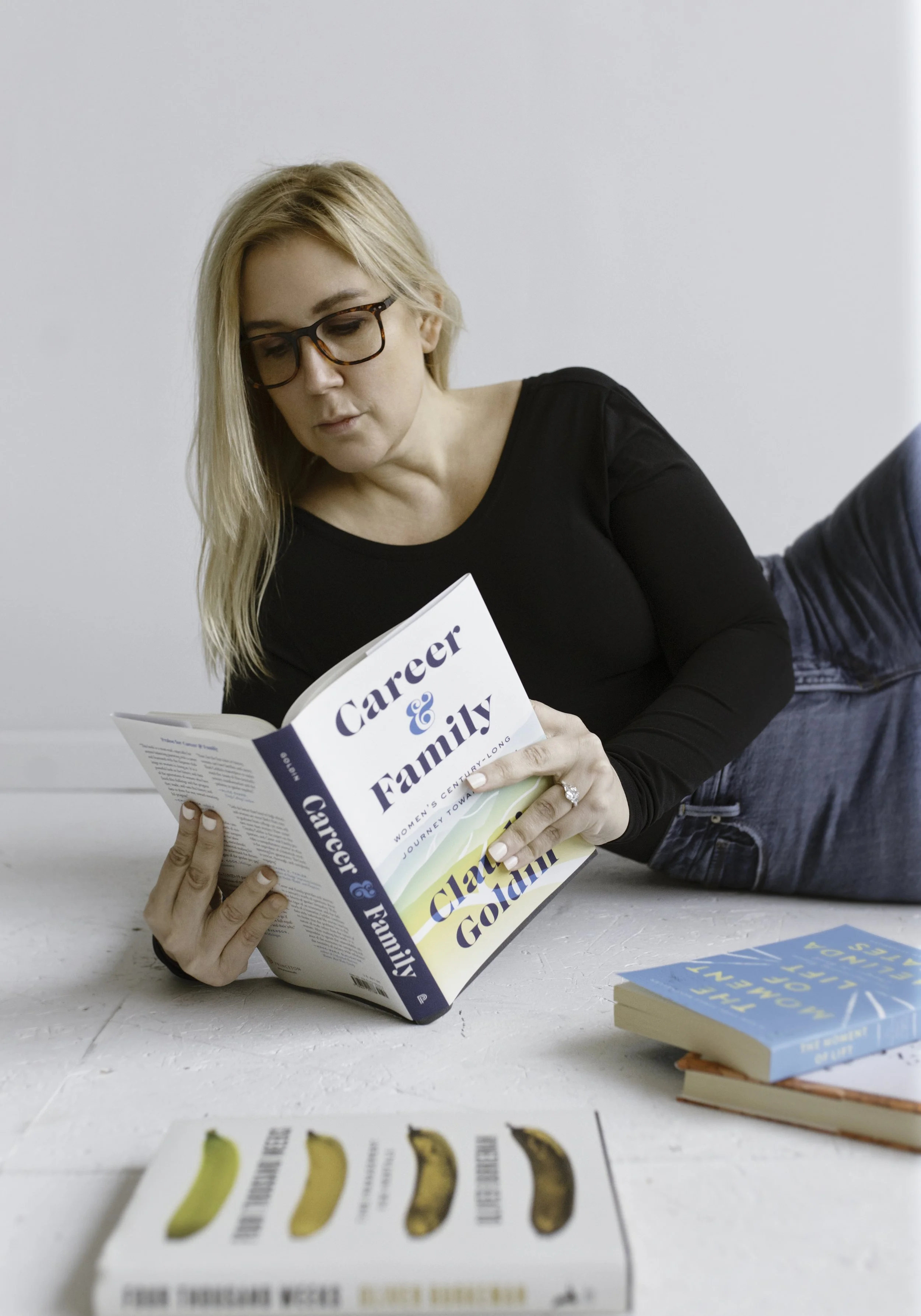 Andrea Mac with blonde hair and glasses is lying on her stomach reading a book titled 'Career & Family' while on a white table. There are several other books and pamphlets on the table.