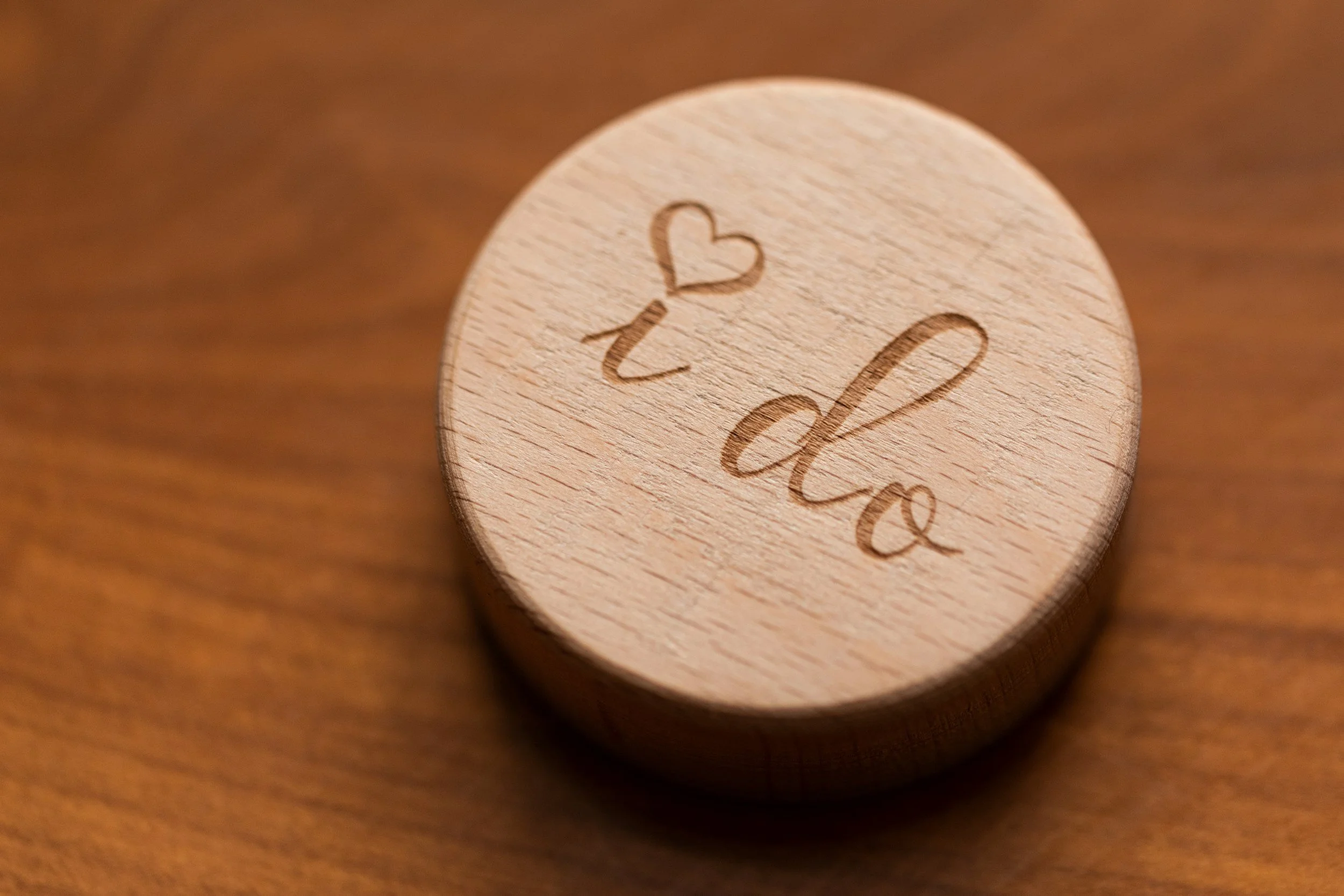 A round wooden disc with the words "I do" and a small heart engraved on its surface, placed on a wooden surface.