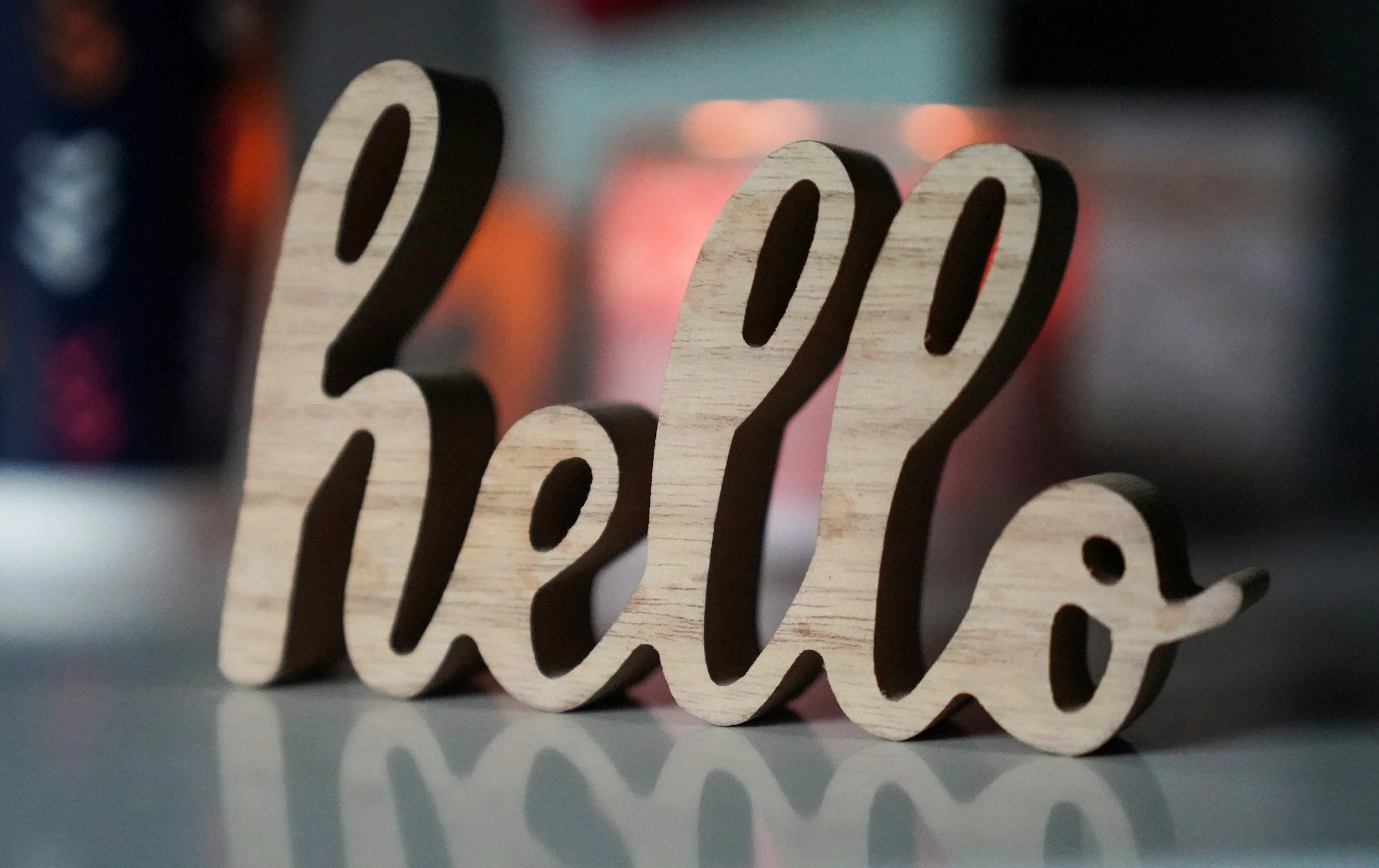 Wooden word cutout spelling "hello" placed on a reflective surface with blurry colorful background.
