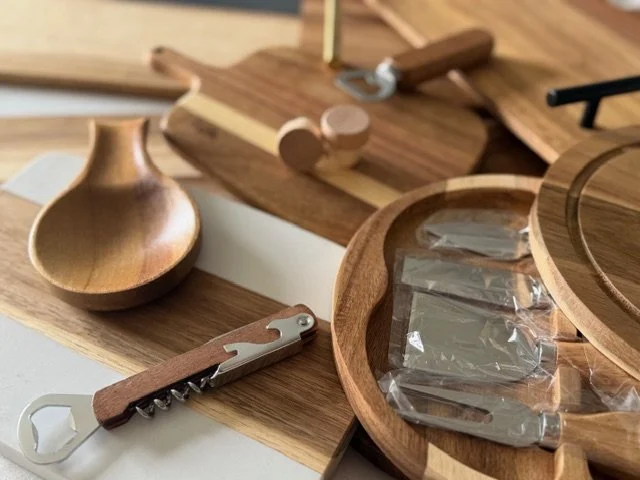 Various wooden kitchen utensils and storage containers on a countertop, including a wooden spoon, a corkscrew wine opener, a round wooden bowl, a rectangular cutting board, and a round wooden container with a transparent lid.