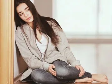 Young woman sitting on the floor resting against a wall, wearing a gray cardigan and jeans.
