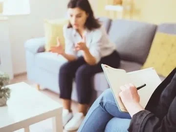 A woman sitting on a sofa during a therapy session, talking and gesturing with her hands while a therapist takes notes.