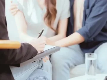 A person in a dark jacket is taking notes on a notepad during a conversation or interview.