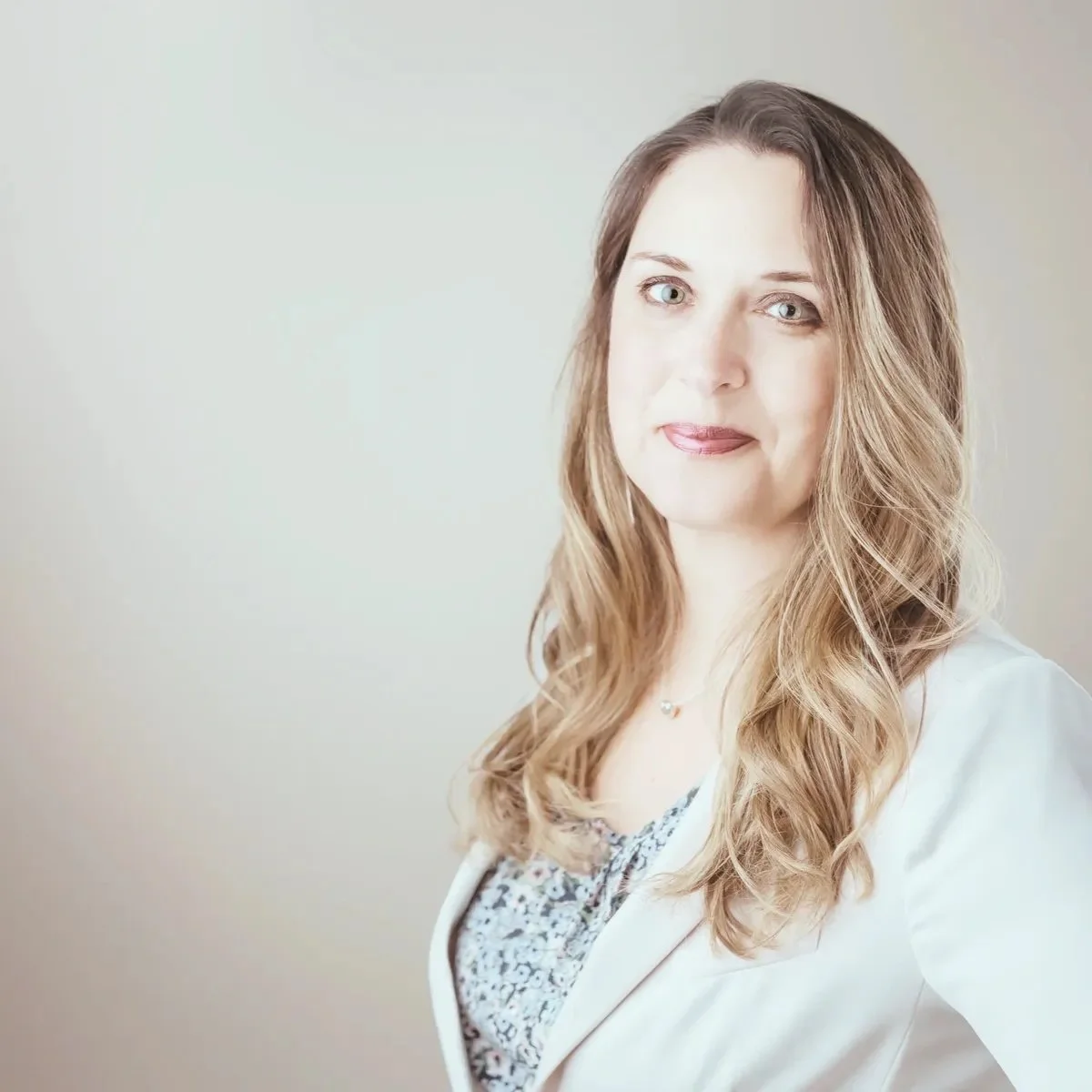 A woman with long, wavy brown hair and light skin, wearing a white blazer over a patterned top, standing against a plain light background.