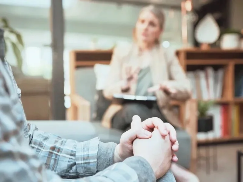 A person holding their hands together during a therapy session with a counselor in the background.