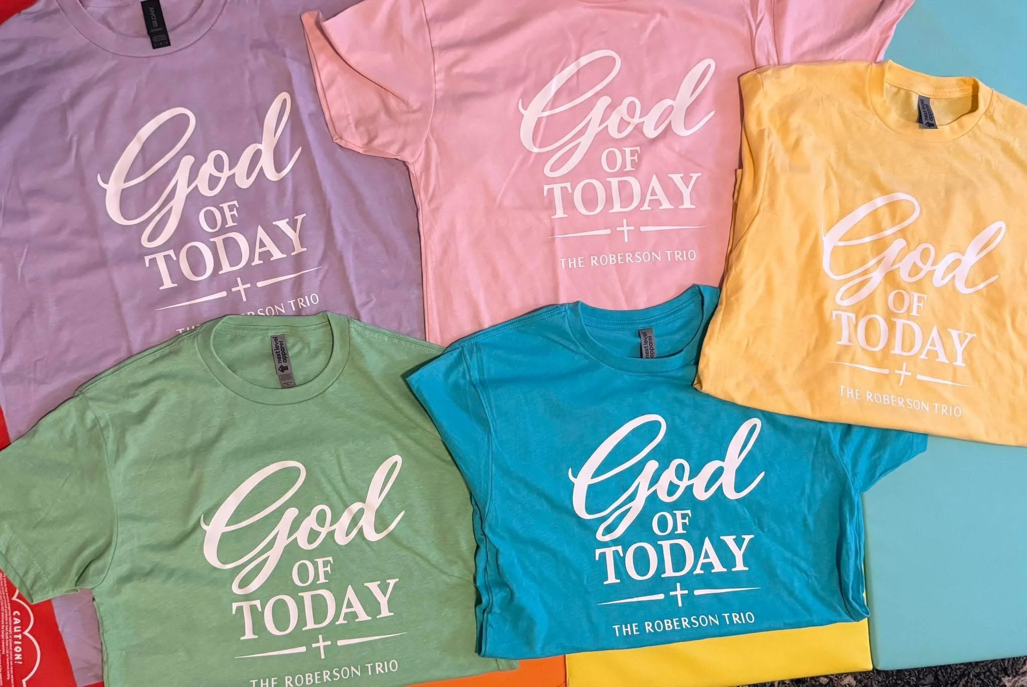 Colorful T-shirts with the phrase 'God of Today' and 'The Roberson Trio' printed on them in white, arranged on a surface.