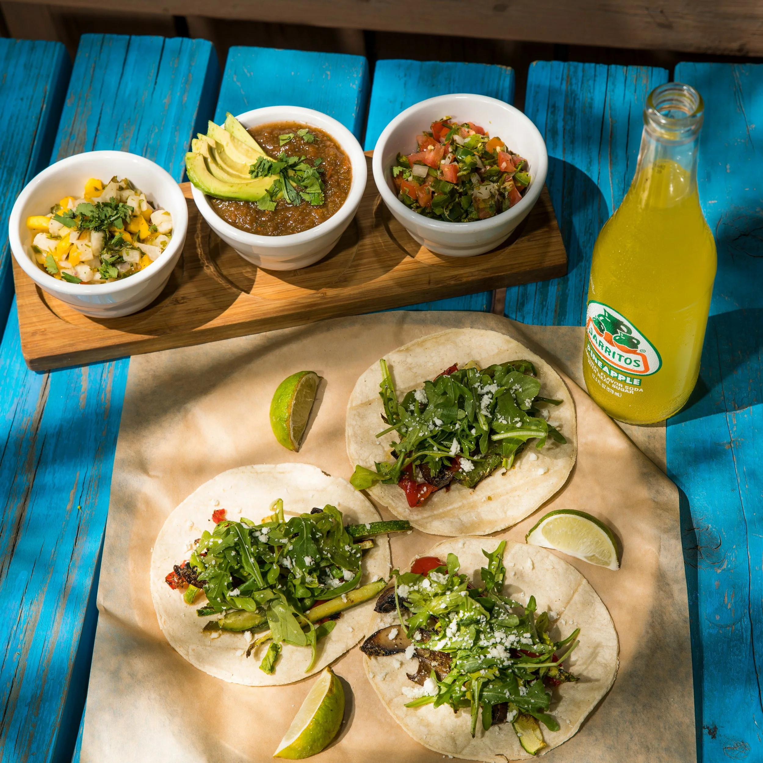 Tacos with greens and cheese on parchment paper, lime wedges, three small bowls with salsa, avocado, and pico de gallo, and a glass bottle of yellow soda on a blue wooden table.