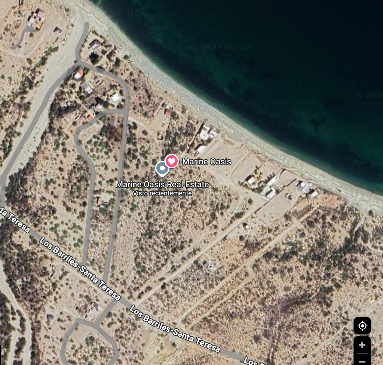 Satellite image showing a coastal residential area with dirt roads, scattered houses, and vegetation near a dark blue ocean or sea.