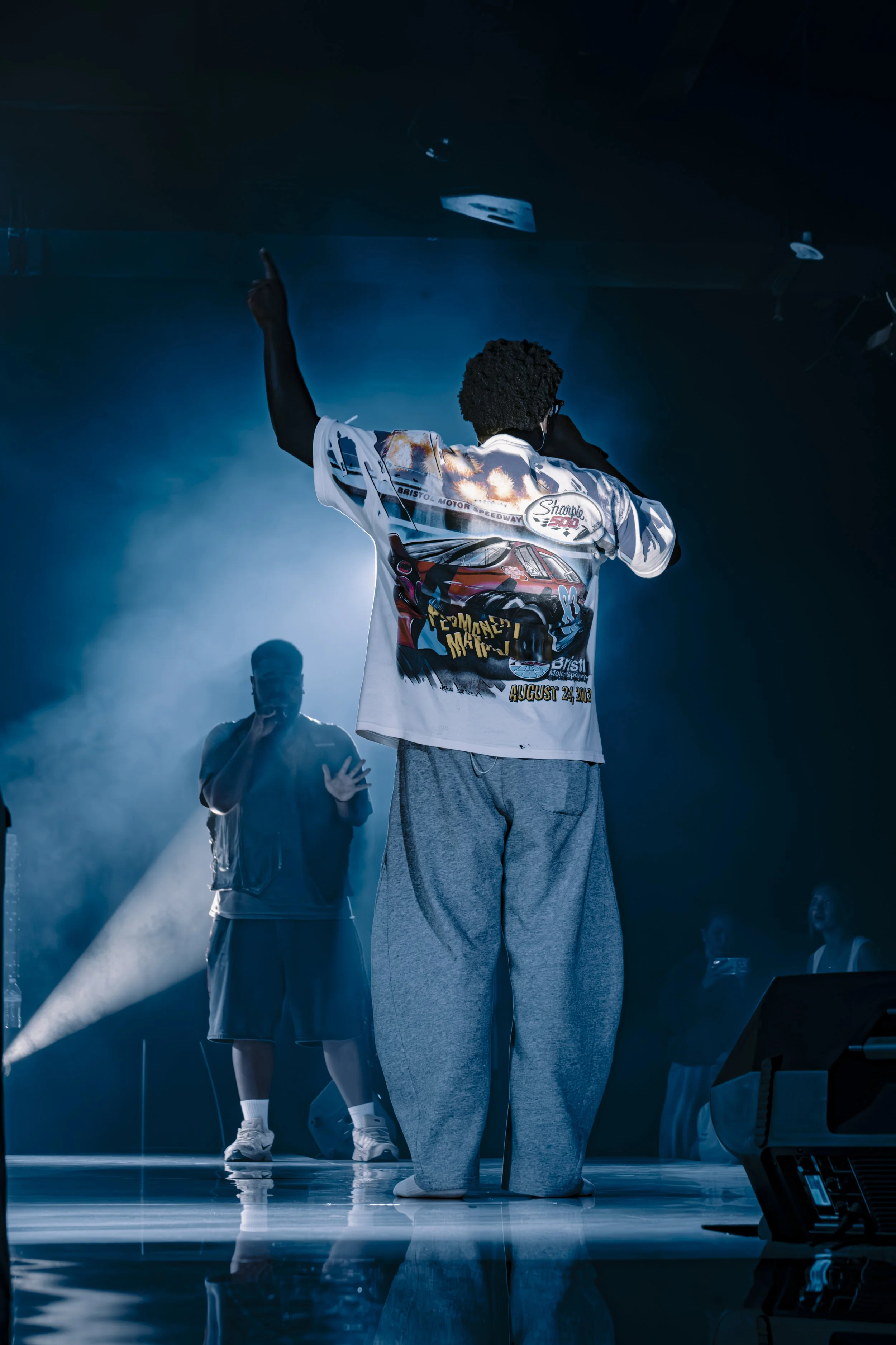 A person with curly hair and glasses on stage, wearing a colorful sports jersey, appears to be singing or speaking into a microphone. Another person in a hoodie stands nearby, and there are stage lights and other people in the background.