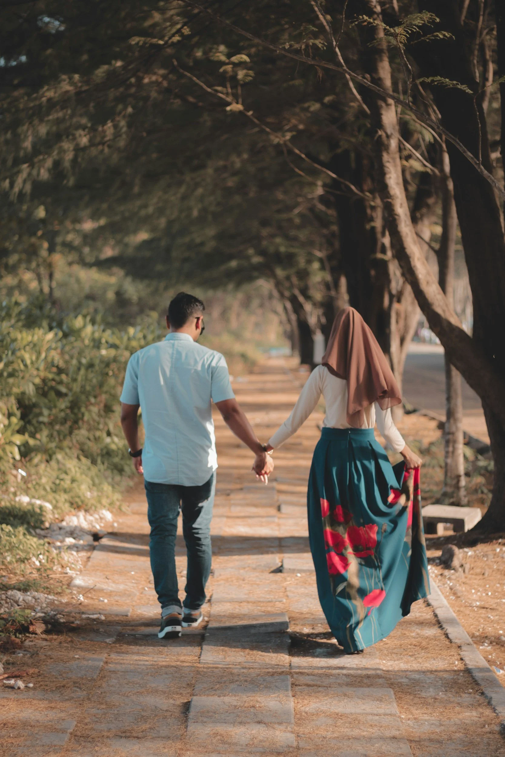 A couple walking hand in hand down a tree-lined pathway on a sunny day, with the woman wearing a long skirt and a headscarf.