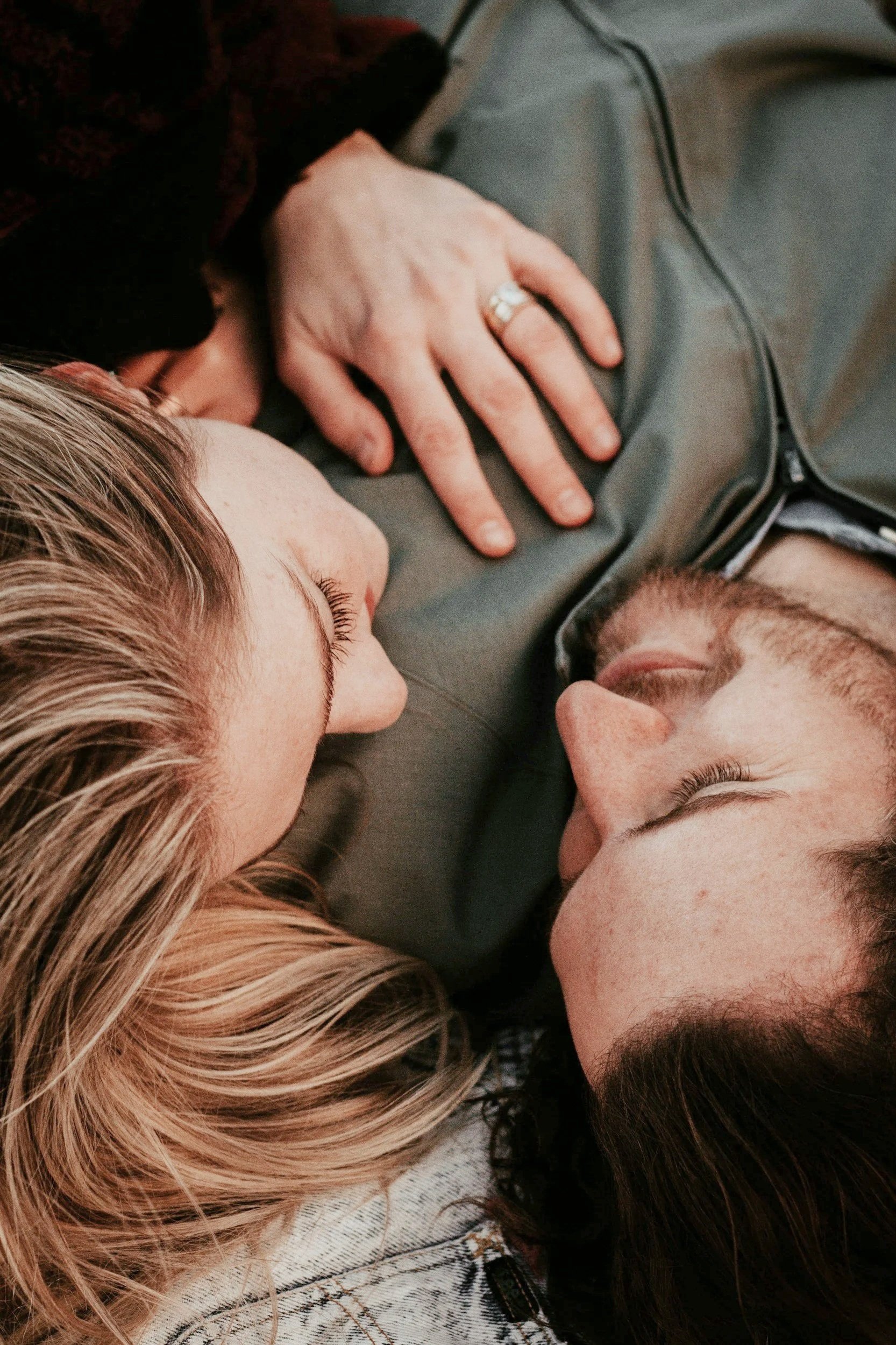 Close-up of two women lying face to face, with one woman's left hand resting on the other woman's chest, showing a ring on her finger. They are lying on a surface, and both have their eyes closed.