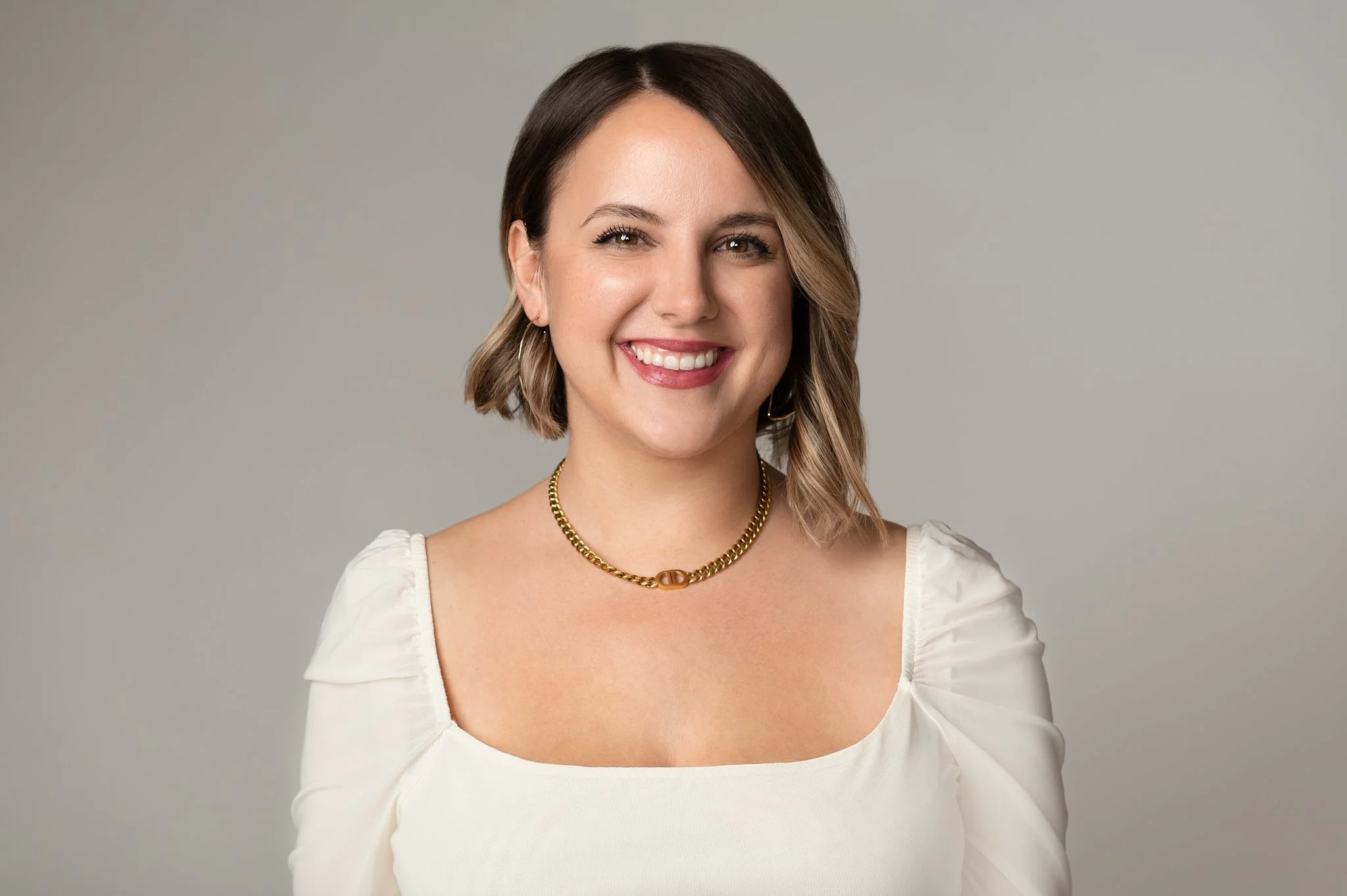 Cat Damare, founder of The Petite Riot. A woman with short, wavy brown hair and light skin smiling, wearing a white top, gold chain necklace, and small hoop earrings, against a plain gray background.