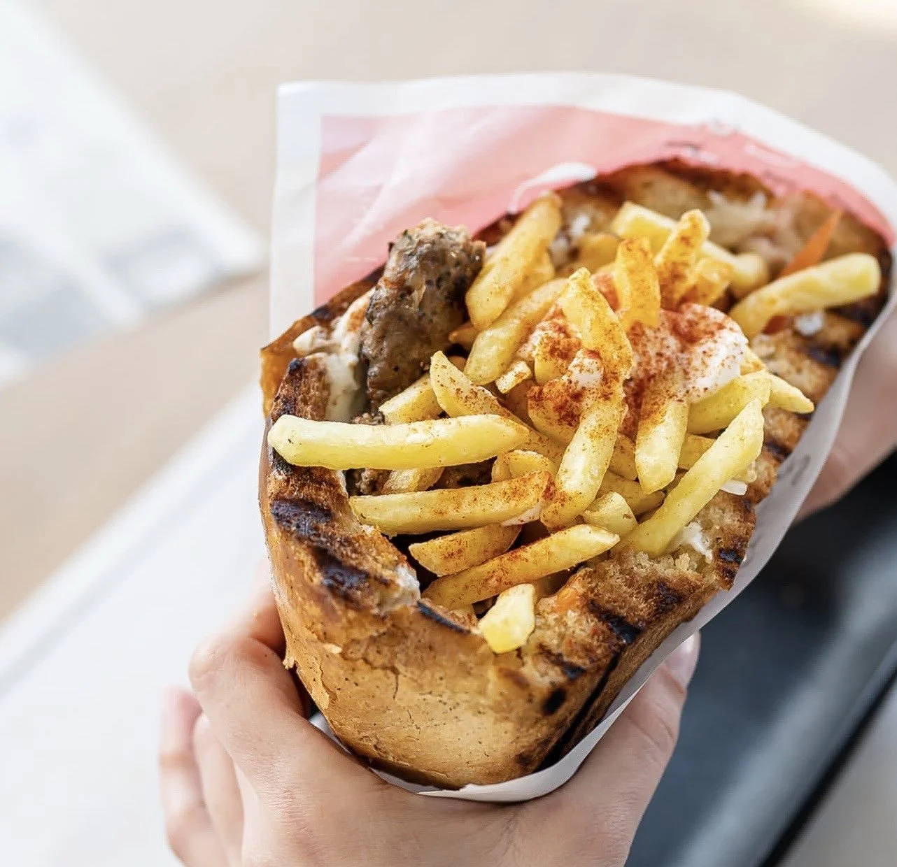 A hand holding a serving of a fast food gyro with French fries in a paper wrapper.