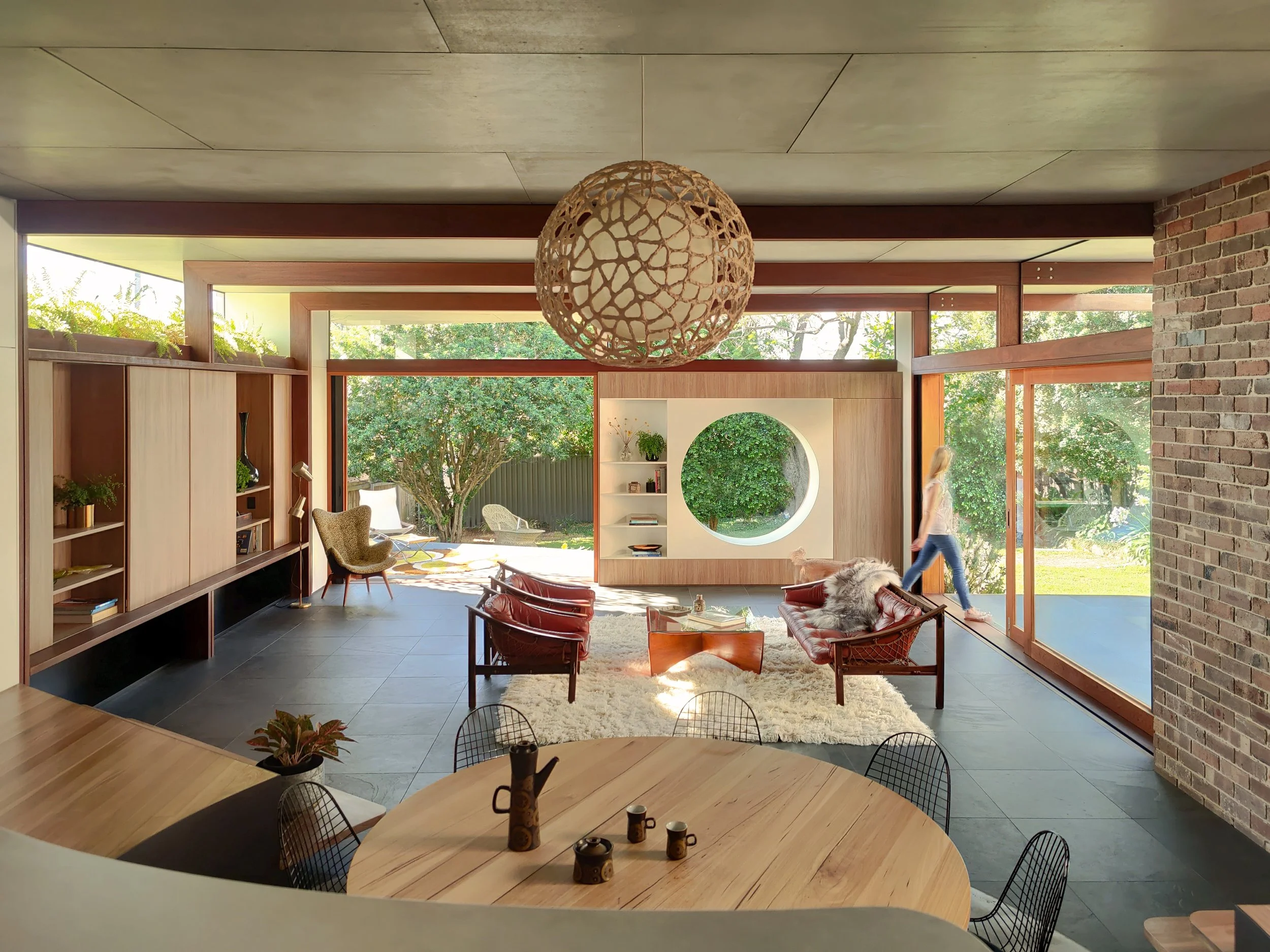 Modern living room with large windows, wooden accents, a round light fixture hanging from the ceiling, and outdoor greenery visible through the windows. Furniture includes chairs, a rug, and shelves with decorative items.