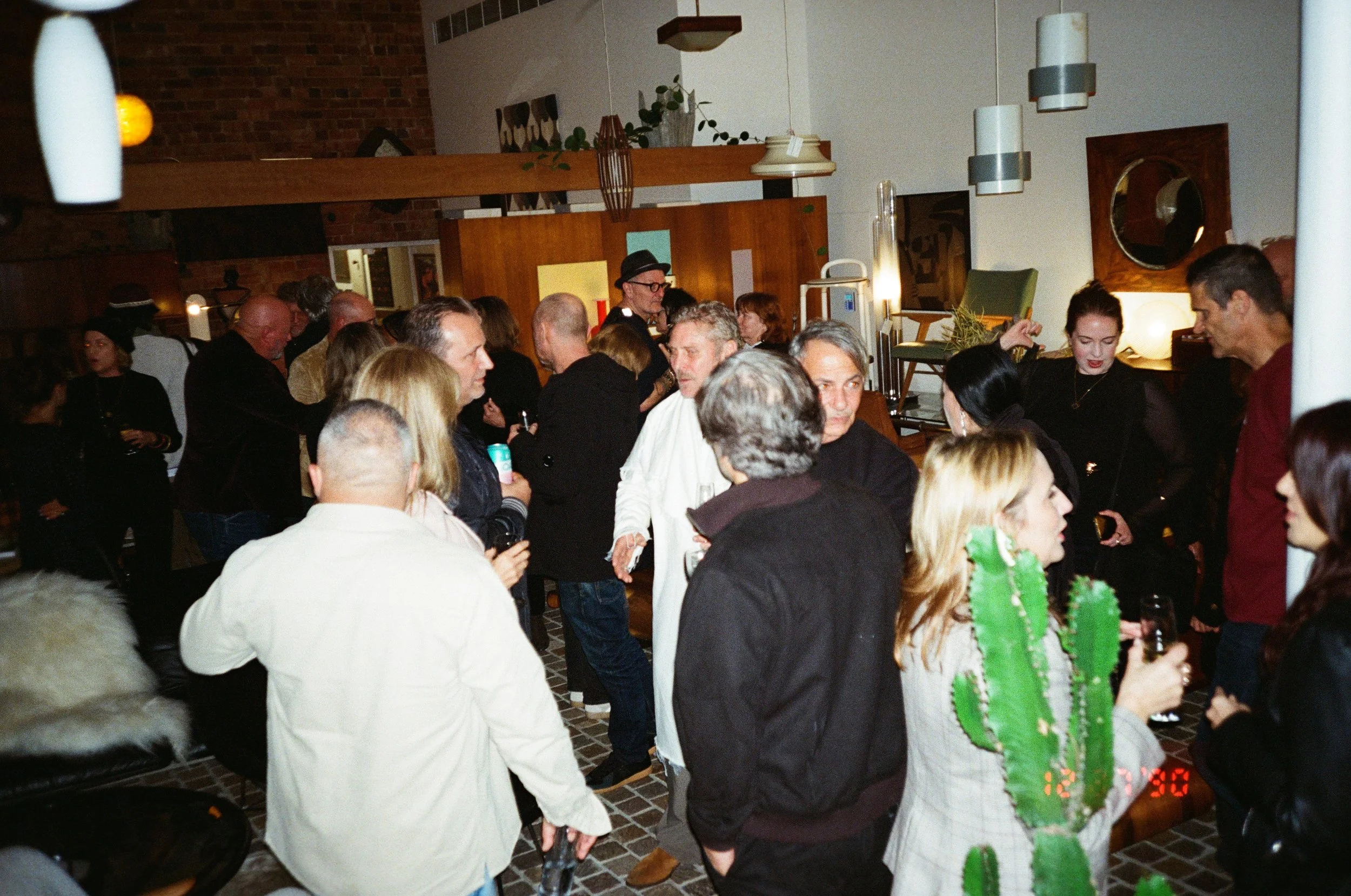 A crowd of people socializing indoors in a warmly lit space with modern decor, including hanging lights and framed wall art.