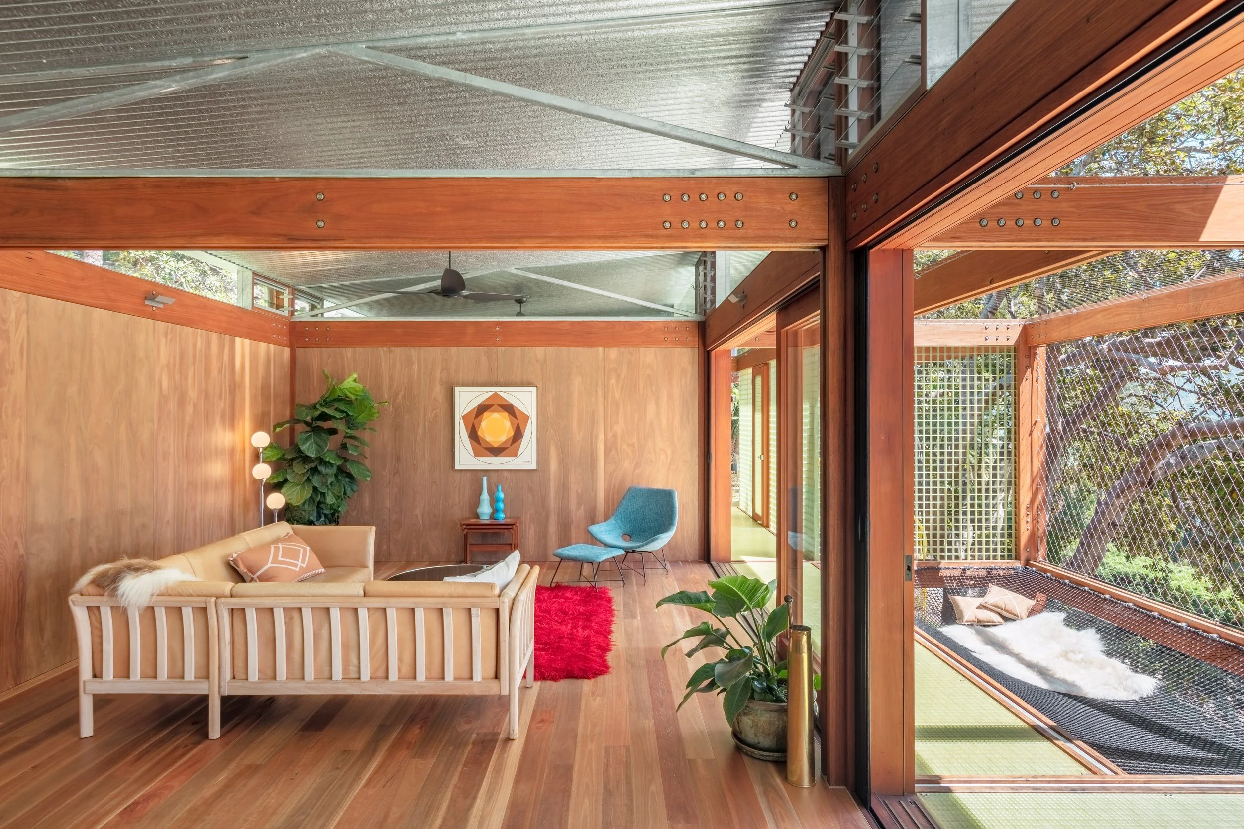 Living room with wooden walls and floors, featuring a beige sofa with pillows, a blue chair, and a small table with vases. Large windows and glass doors reveal an outdoor area with trees and a screened porch.
