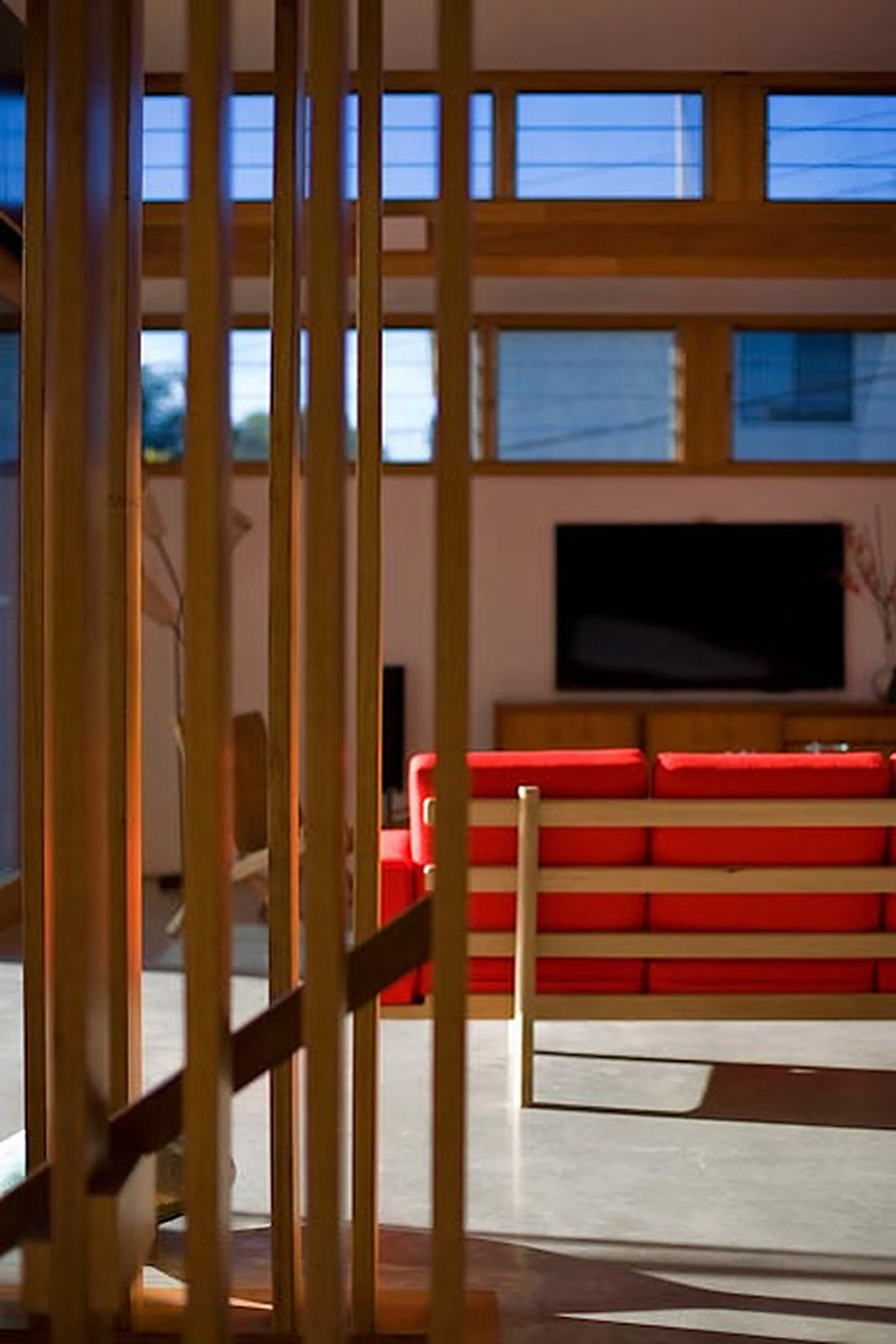View of a modern living room through wooden slats, featuring a red and beige striped sofa, a flat-screen TV on the wall, and large windows with blinds letting in natural light.