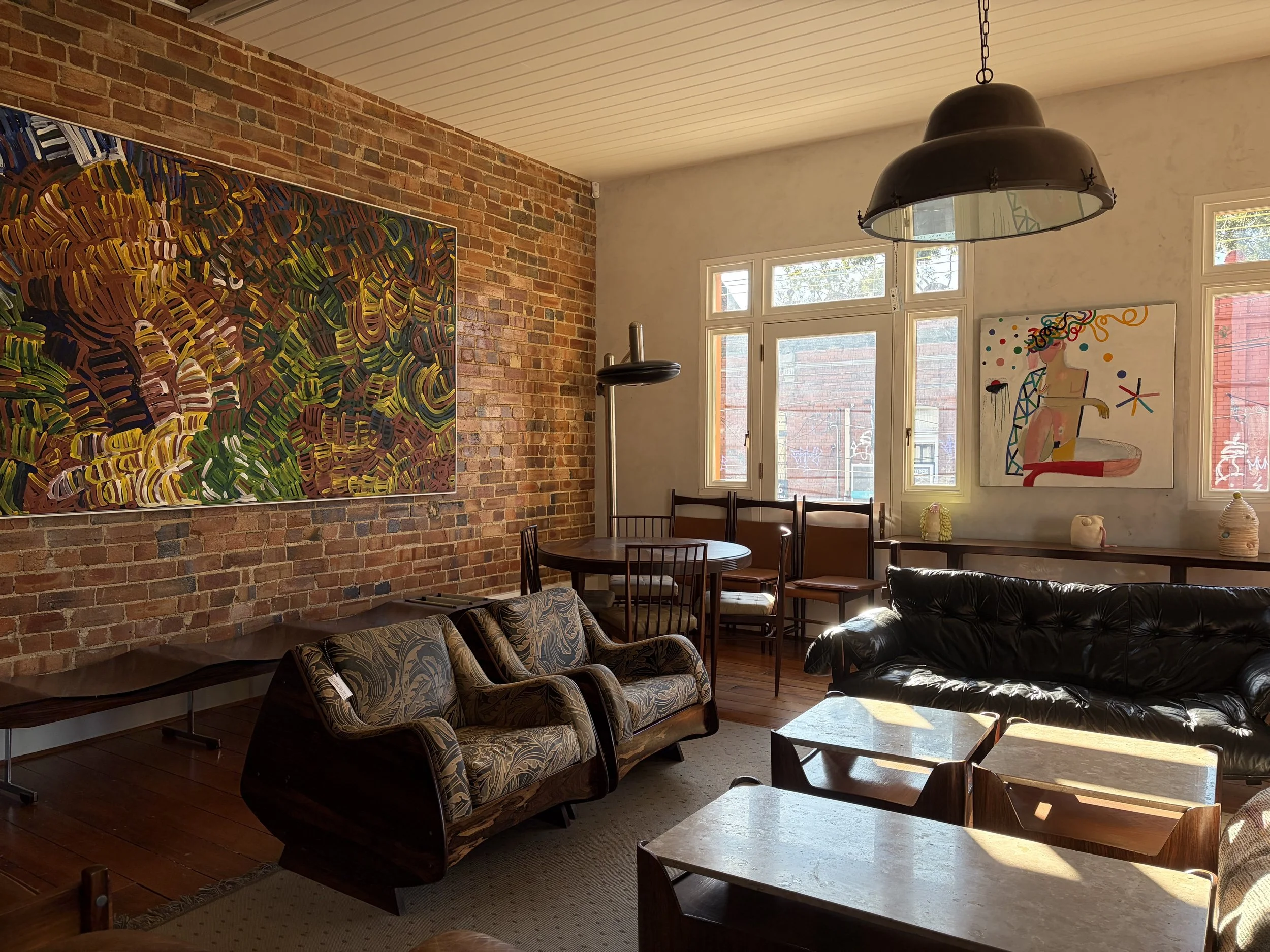 A cozy room with exposed brick wall and white painted ceiling. Large windows let in natural light. Decor includes colorful abstract paintings, vintage-style chairs, and a black leather sofa.
