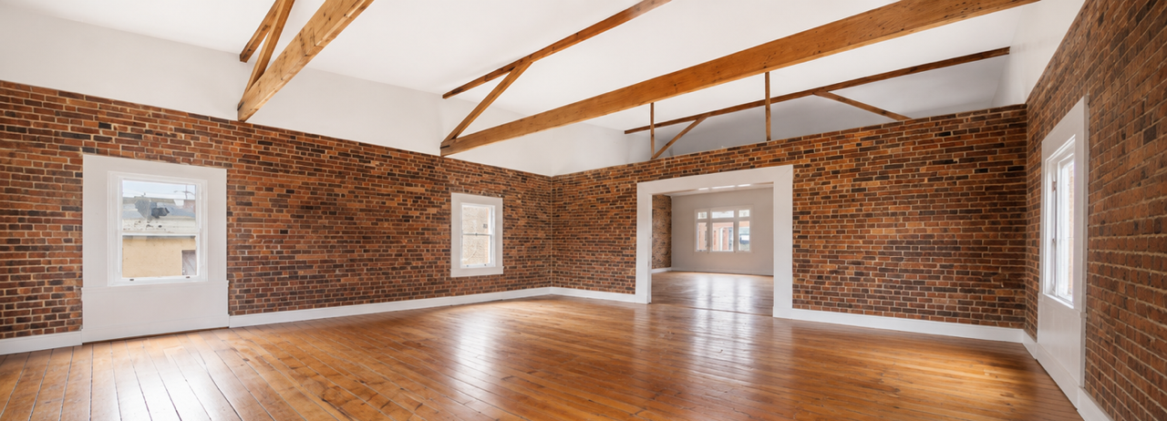 Empty room with brick walls, wooden floors, and exposed wooden beams on the ceiling, with multiple windows providing natural light.