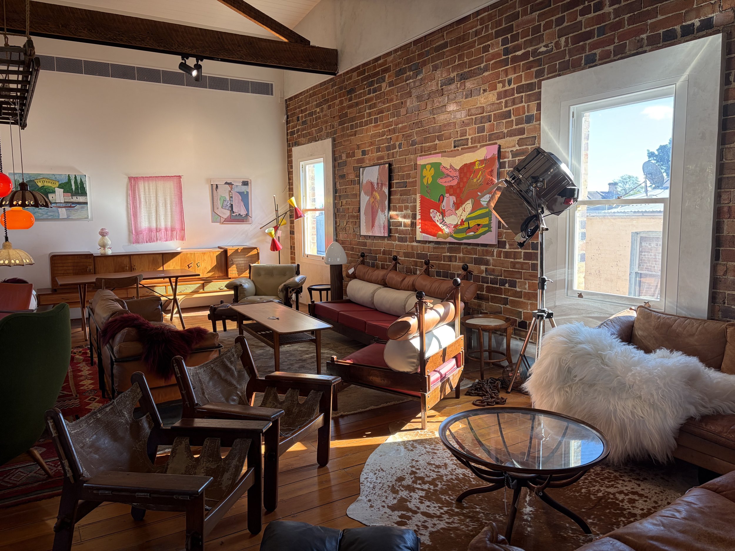 A cozy living room with a brick wall featuring colorful abstract artwork, a large window letting in natural light, a mixture of vintage and modern furniture including chairs, a sofa, and a cowhide rug.