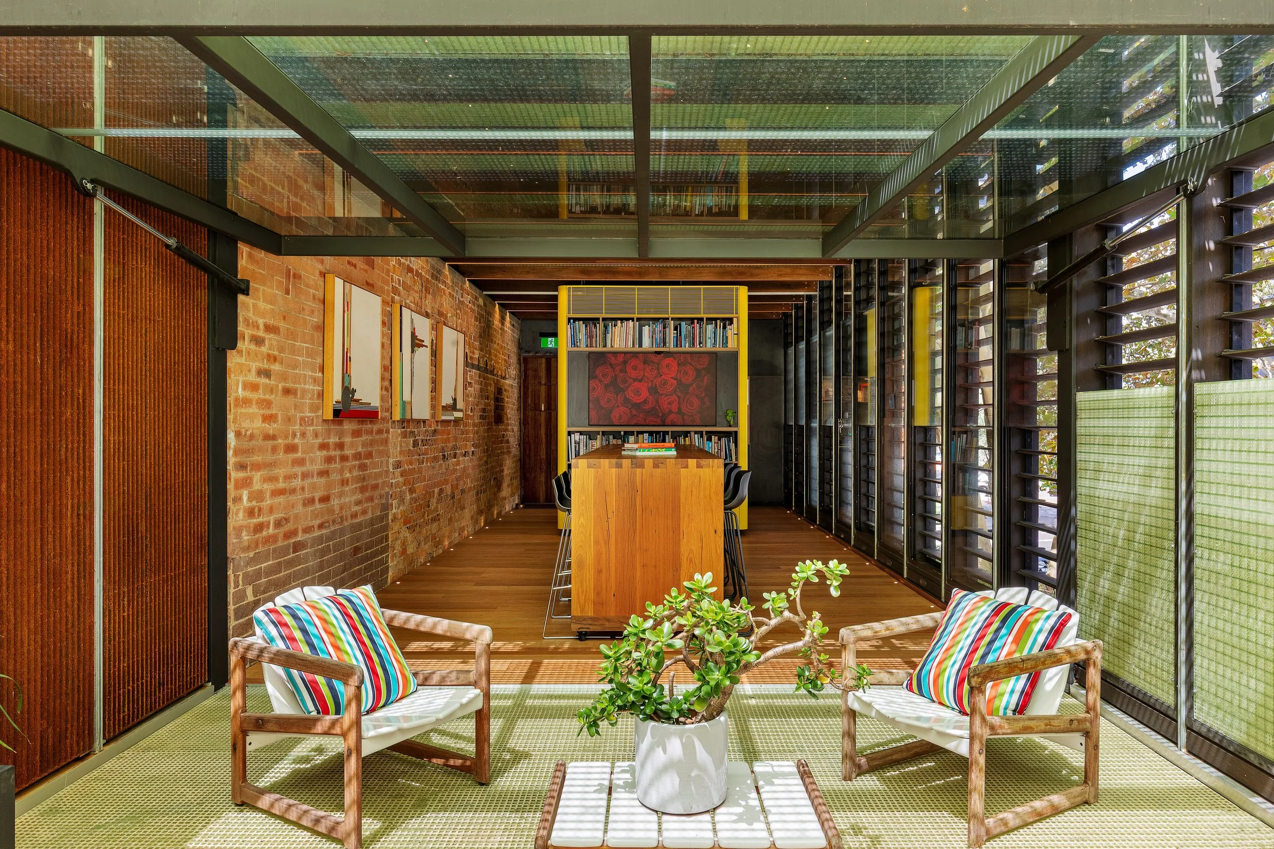 Indoor seating area with two armchairs with colorful striped cushions and a small white table with a potted succulent plant, brick walls, a yellow bookshelf, and large windows with black frames.