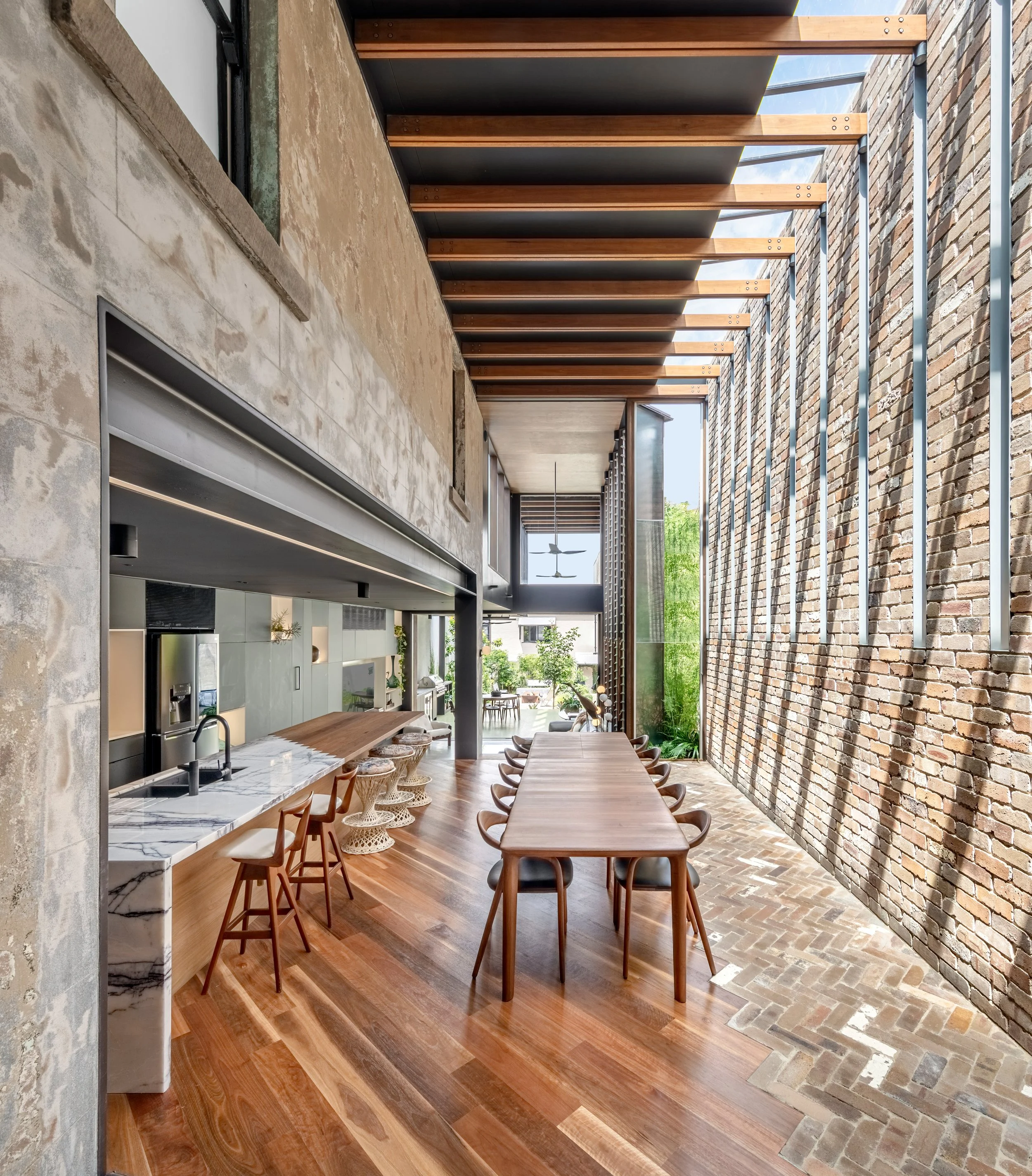 Modern open-plan kitchen and dining area with large window, brick wall, and solar shading structures outside.