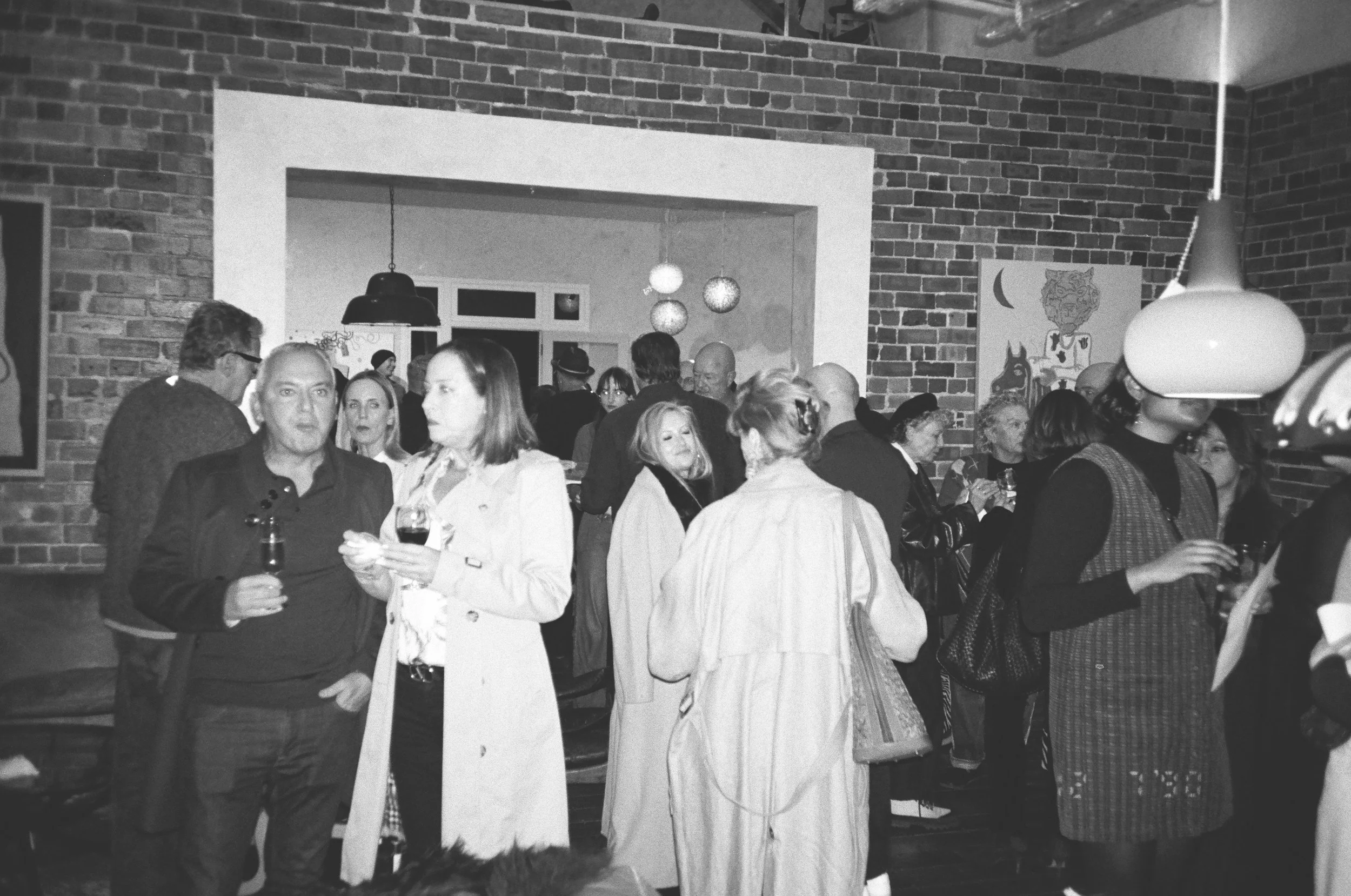 A group of people socializing at an indoor gathering, with brick walls, pendant lighting, and artwork on the walls, some holding drinks and engaging in conversation.
