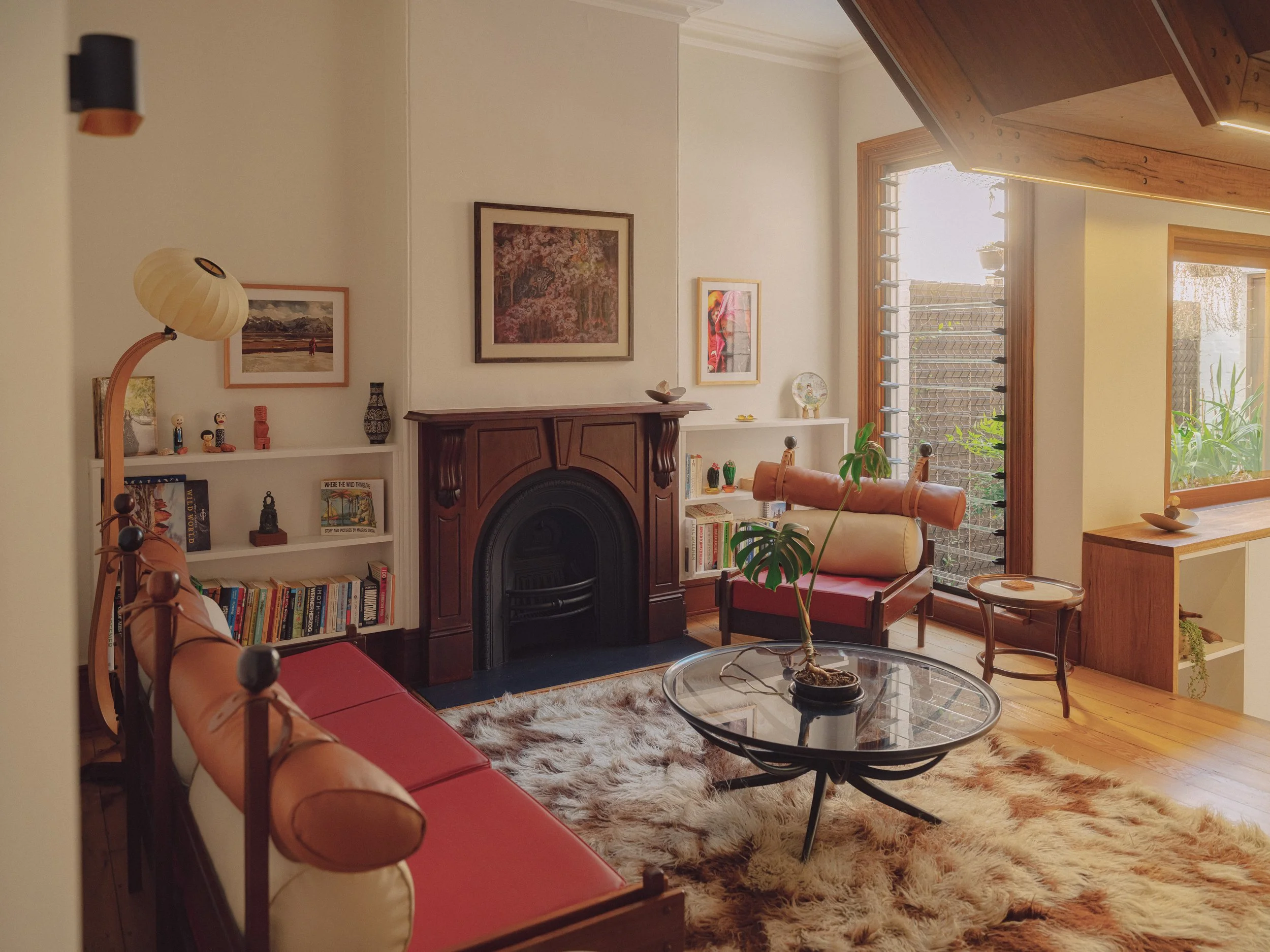 A cozy living room with wooden flooring, a red couch, a glass coffee table with a potted plant, a rug, and a fireplace. There are framed pictures and artwork on the walls, shelves with books and decorative items, and large windows with wooden frames letting in natural light.