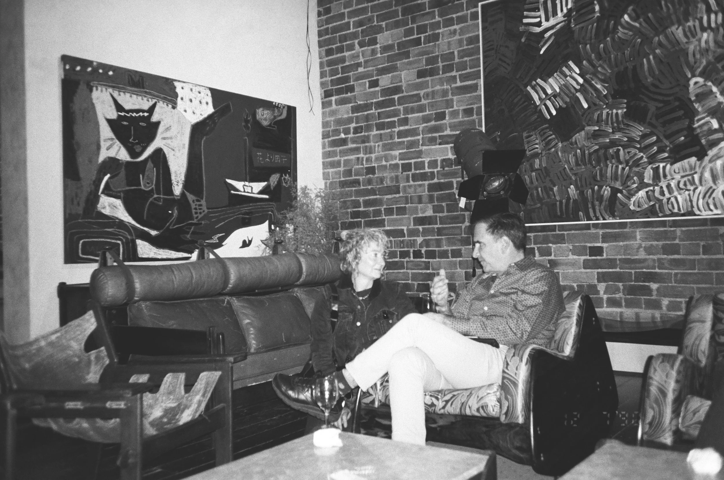 A black-and-white photo of a man and a woman sitting on a couch in a brick-walled room, engaging in conversation. Artworks decorate the wall behind them.